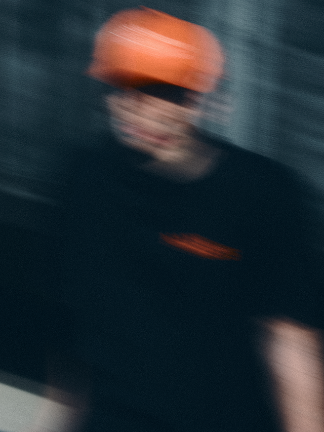 Model in schwarzem NAYS College Print T-Shirt und orangener Fishing Club Cap im urbanen Setting des Velodrom Berlin. Verwackeltes Foto.