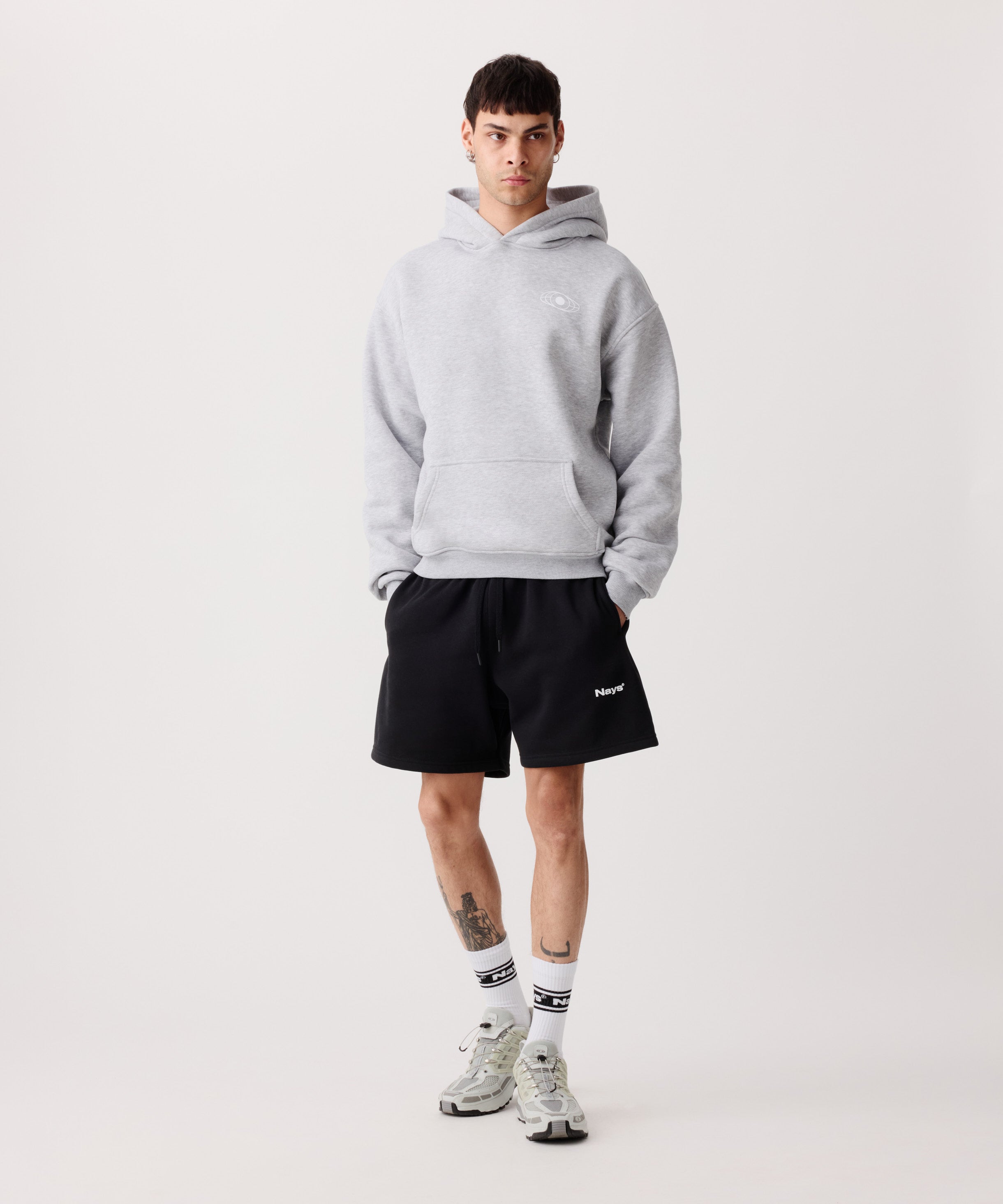 Ein hell-grauer Kapuzen-Pullover getragen von einem Mann. Er trägt dazu 
eine kurze, schwarze Hose, weiße Socken und helle Turnschuhe. Beide Hände sind in den Hosentaschen.