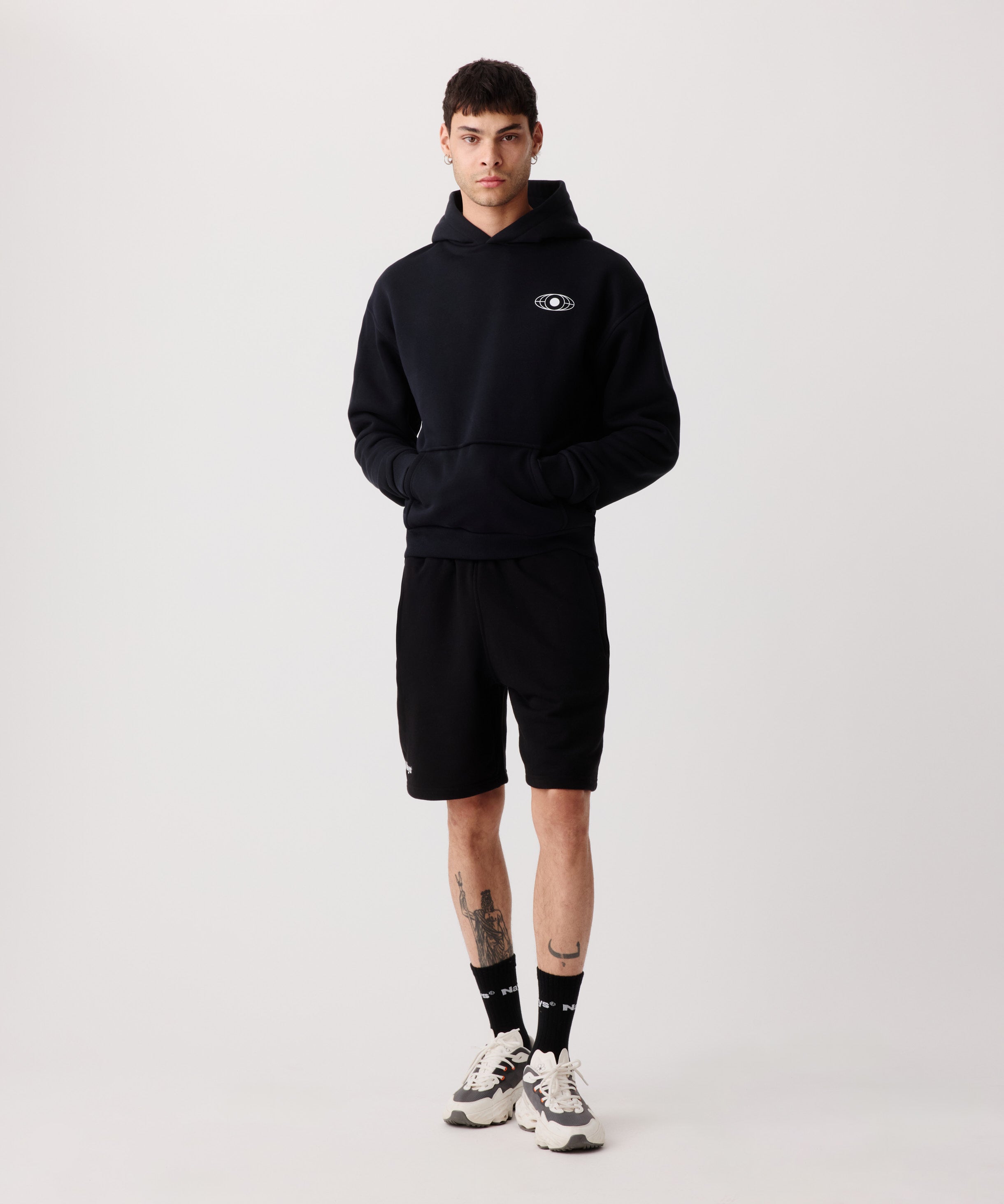 Ein schwarzer Kapuzen-Pullover getragen von einem Mann. Er trägt dazu 
eine kurze, schwarze Hose, schwarze Socken und helle Turnschuhe. Beide Hände sind in der Känguru-Tasche.