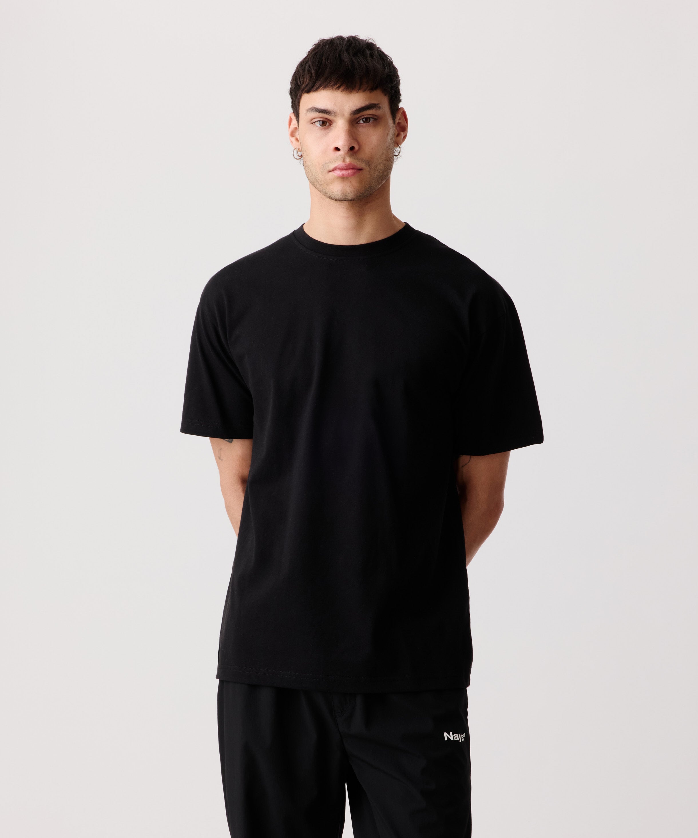 Ein Mann trägt das weiße Logo-T-Shirt mit verschränkten Armen auf dem Rücken. Dazu kombiniert er eine schwarze Hose mit weißem „Nays“-Logo-Schriftzug.