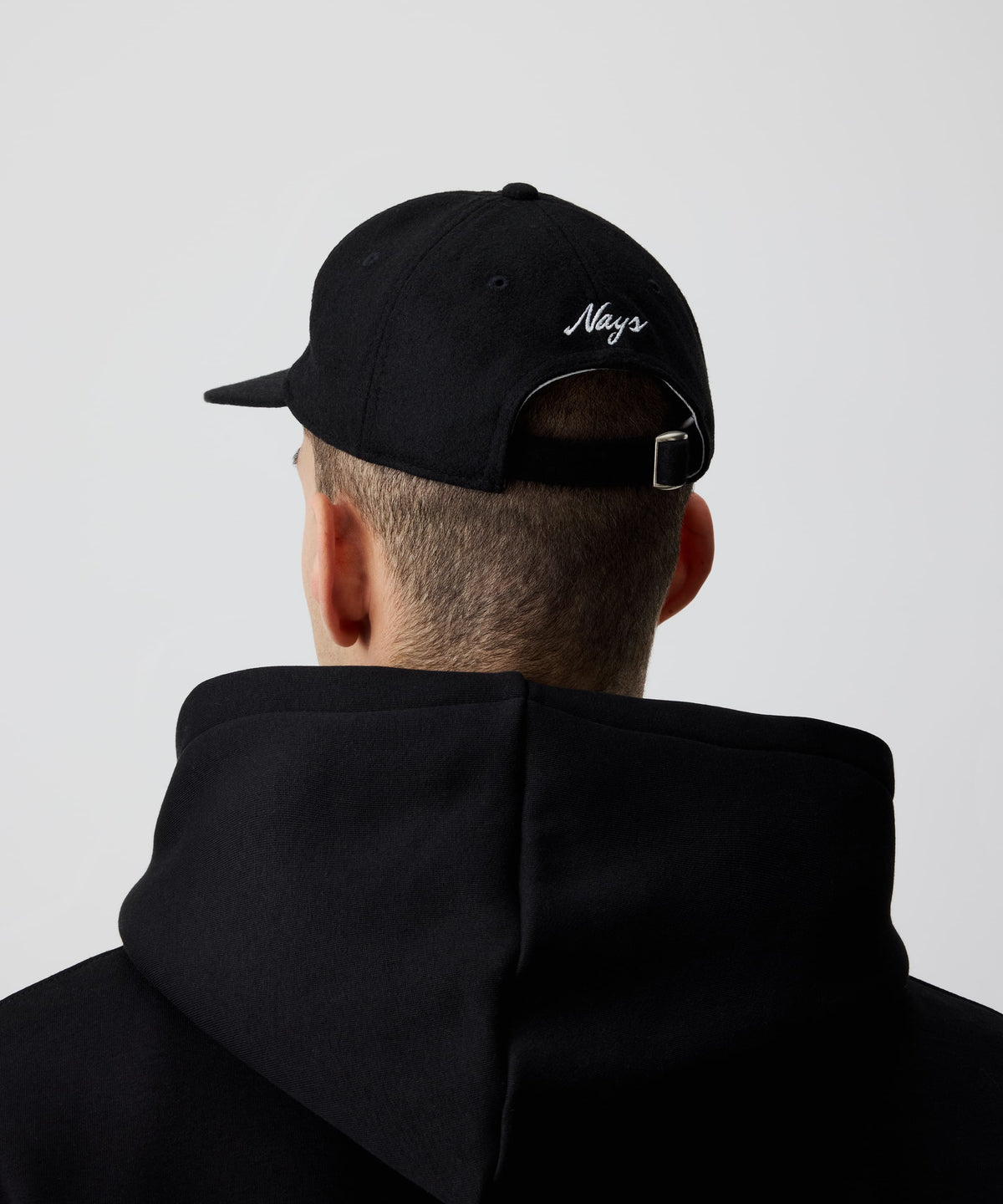 Mann trägt schwarze Nays Felt Cap, eine Schirmmütze aus Filz kombiniert mit einem schwarzen Kapuzen-Pullover in der Rückansicht. Zu erkennen sind das weiße Logo und der größen-verstellbare Riemen.