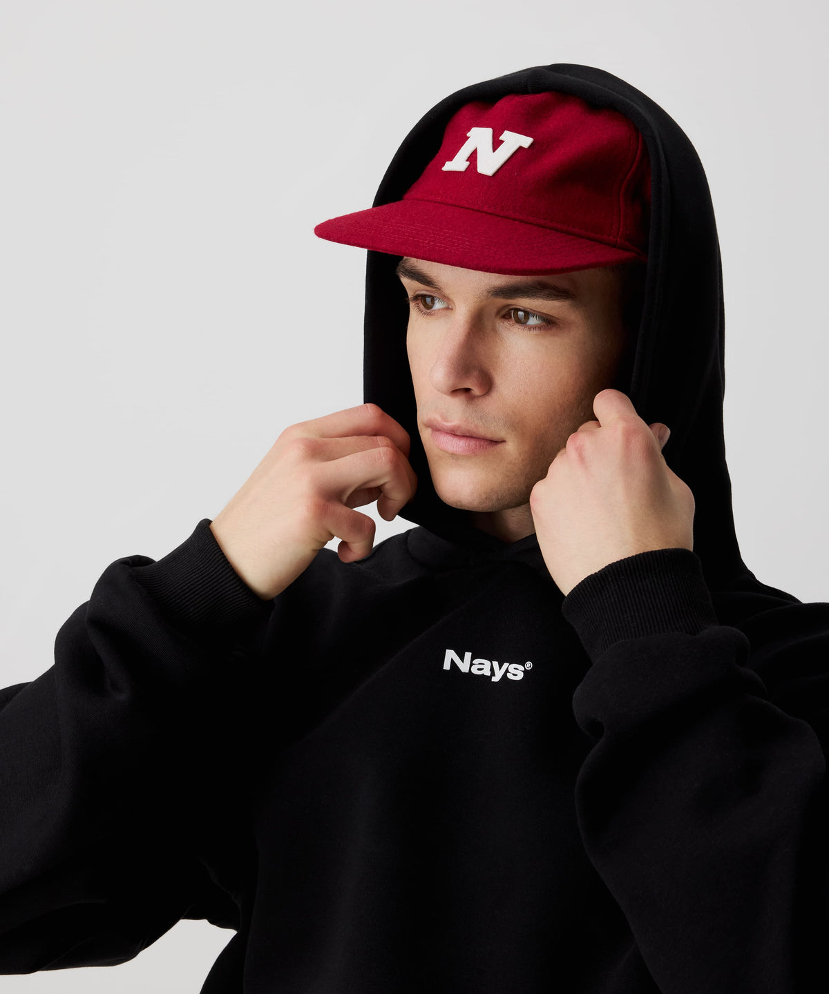 Mann trägt rote Nays Felt Cap, eine stilvolle, robuste Filzmütze kombiniert mit einem schwarzen Kapuzen-Pullover mit weißem Nays-Logo-Print auf der Brust.