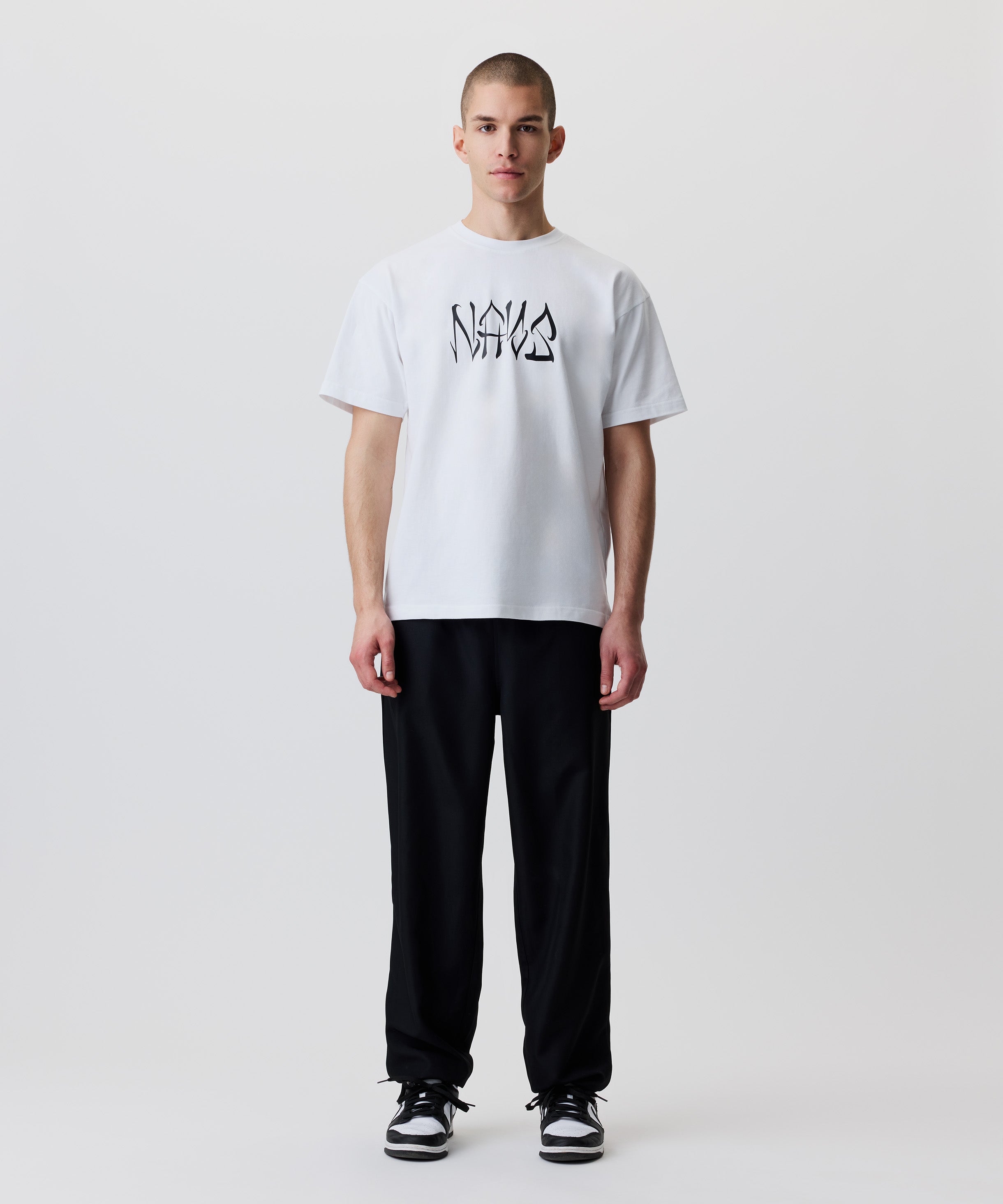 Das Bild zeigt die schwarze Hose getragen von einem Mann. Er trägt dazu ein weißes T-Shirt mit schwarzem Logo und schwarz-weiße Turnschuhe.