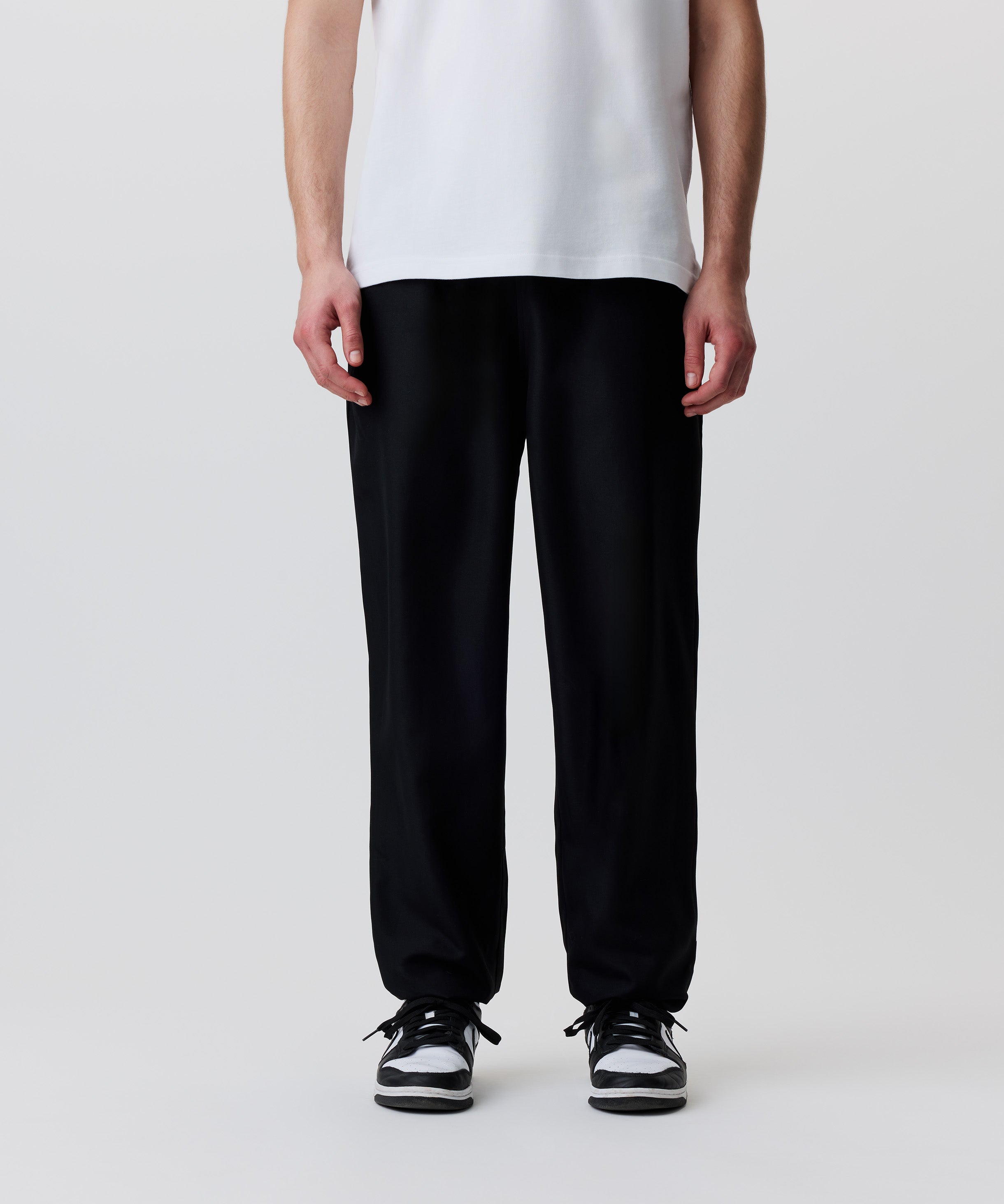 Der Bild-Ausschnitt zeigt die schwarze Hose getragen von einem Mann. Er trägt dazu ein weißes T-Shirt und schwarz-weiße Turnschuhe.