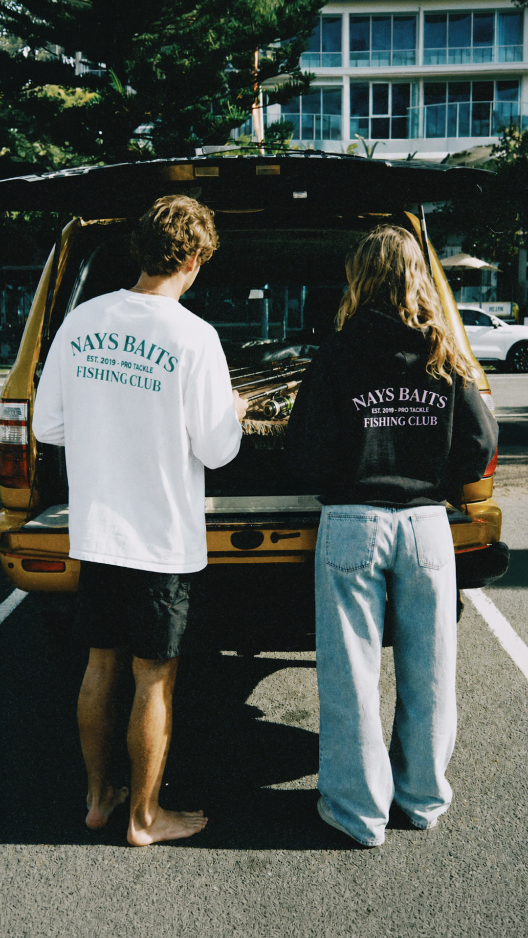 Zwei Personen in Nays Baits Fishing Club Streetwear vor einem Auto mit Angelausrüstung.