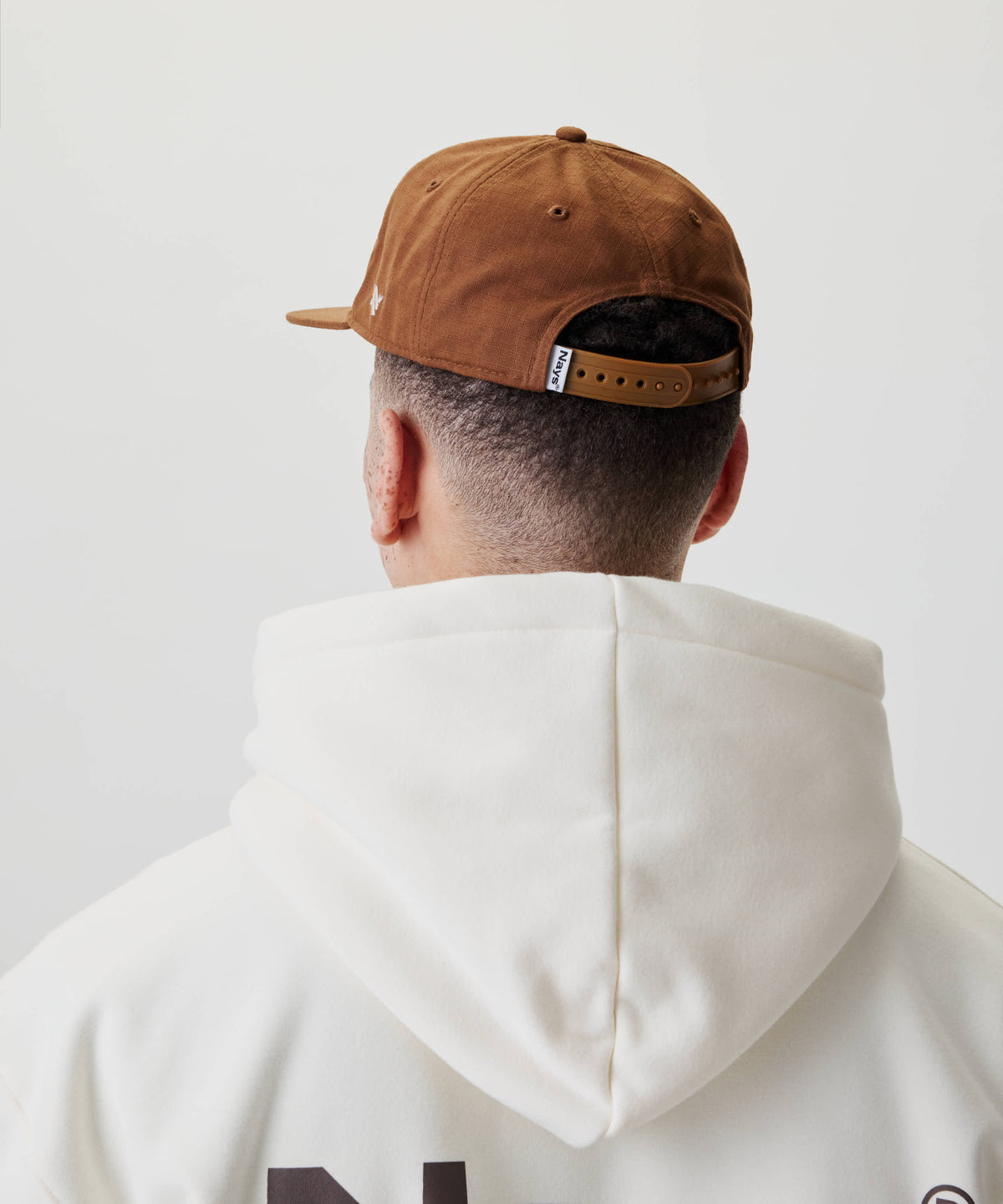Person von hinten mit einem nach hinten gedrehten Ripstop Script Cap mit verstellbarem Riemen, kombiniert mit einem cremefarbenen Hoodie ohne Kapuze.