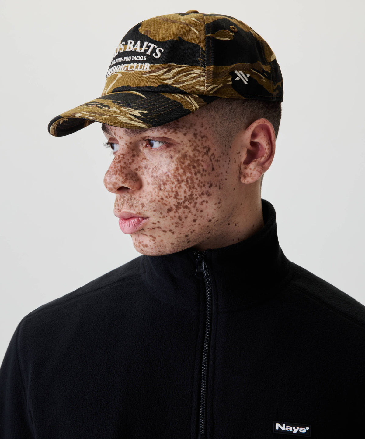 Junger Mann mit Nays Fishing Club Cap im markanten Camo-Print, zeigt das 5-Panel-Design und die Off-white Stickerei, getragen mit schwarzem Fleece-Top.