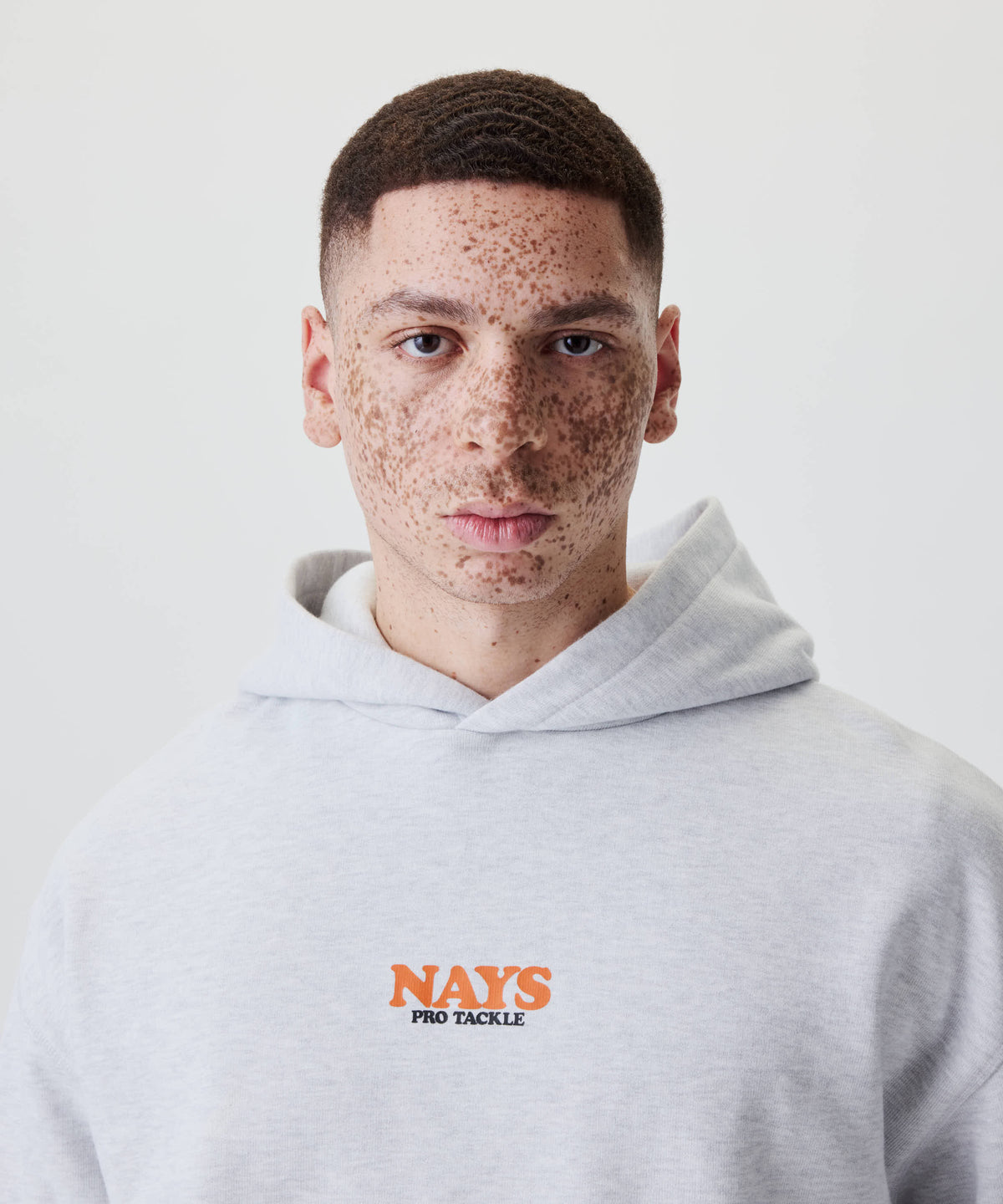Junger Mann mit Sommersprossen trägt den Nays College Print Hoodie in Grau Melange mit auffälligem NAYS PRO TACKLE-Print auf der Brust.