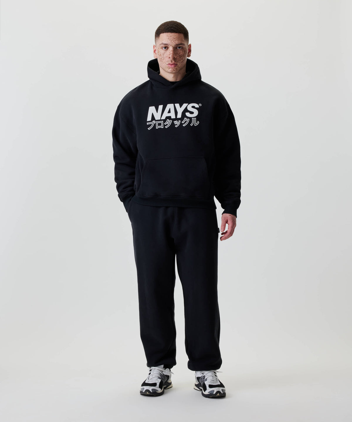 Mann trägt den Nays Understatement Hoodie, schwarz mit weißem Print und japanischen Zeichen, in entspannter Passform, kombiniert mit schwarzen Jogginghosen und sportlichen Sneakern.