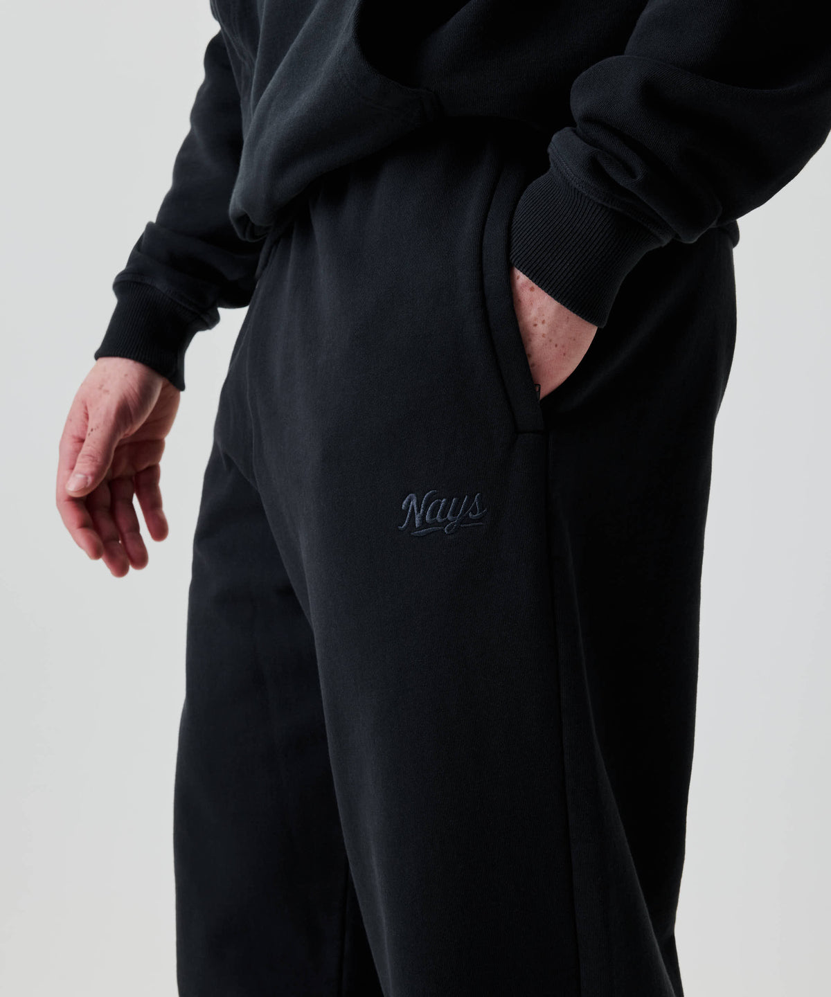 Eine Person trägt Nays Straight Leg Sweatpants mit geradem Bein, seitlichen Taschen und gesticktem Logo Nays auf dem Oberschenkel; eine Hand steckt in der Tasche.
