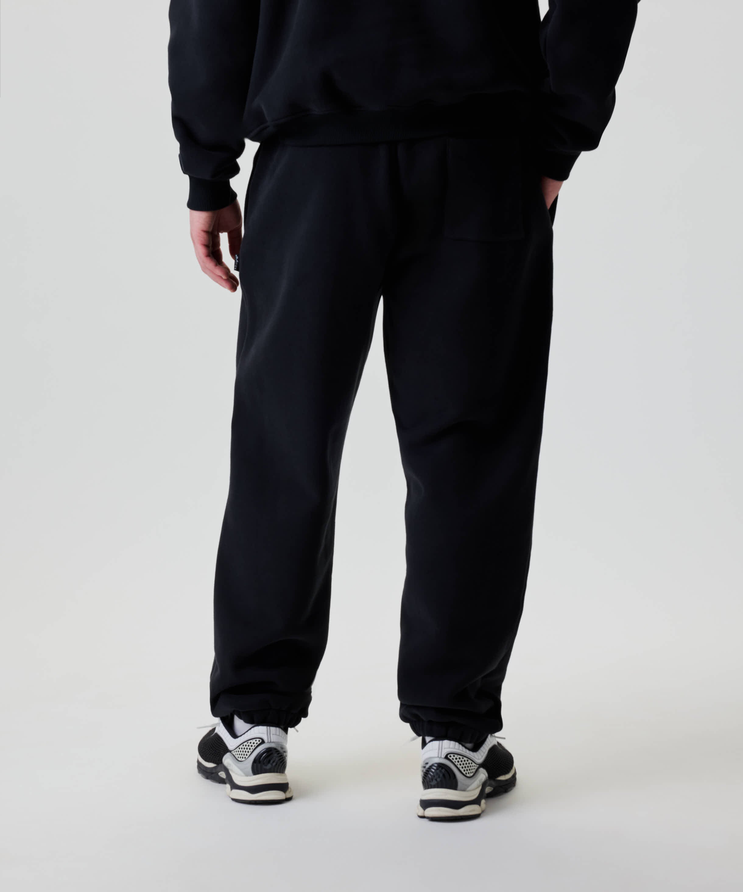 Person trägt Nays Baggy Sweatpants mit elastischen Bündchen und einer rechten Gesäßtasche, kombiniert mit schwarzen Sneakers, linke Hand in der Tasche.