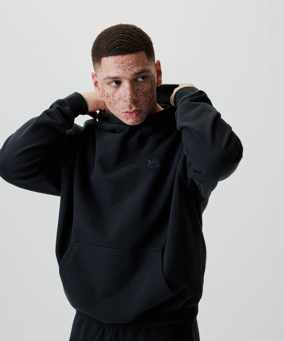 Junger Mann trägt den Nays New Script Hoodie mit Logo-Stickerei. Er hält die Kapuze hinten, Blick zur Seite, minimalistischer Studio-Look.