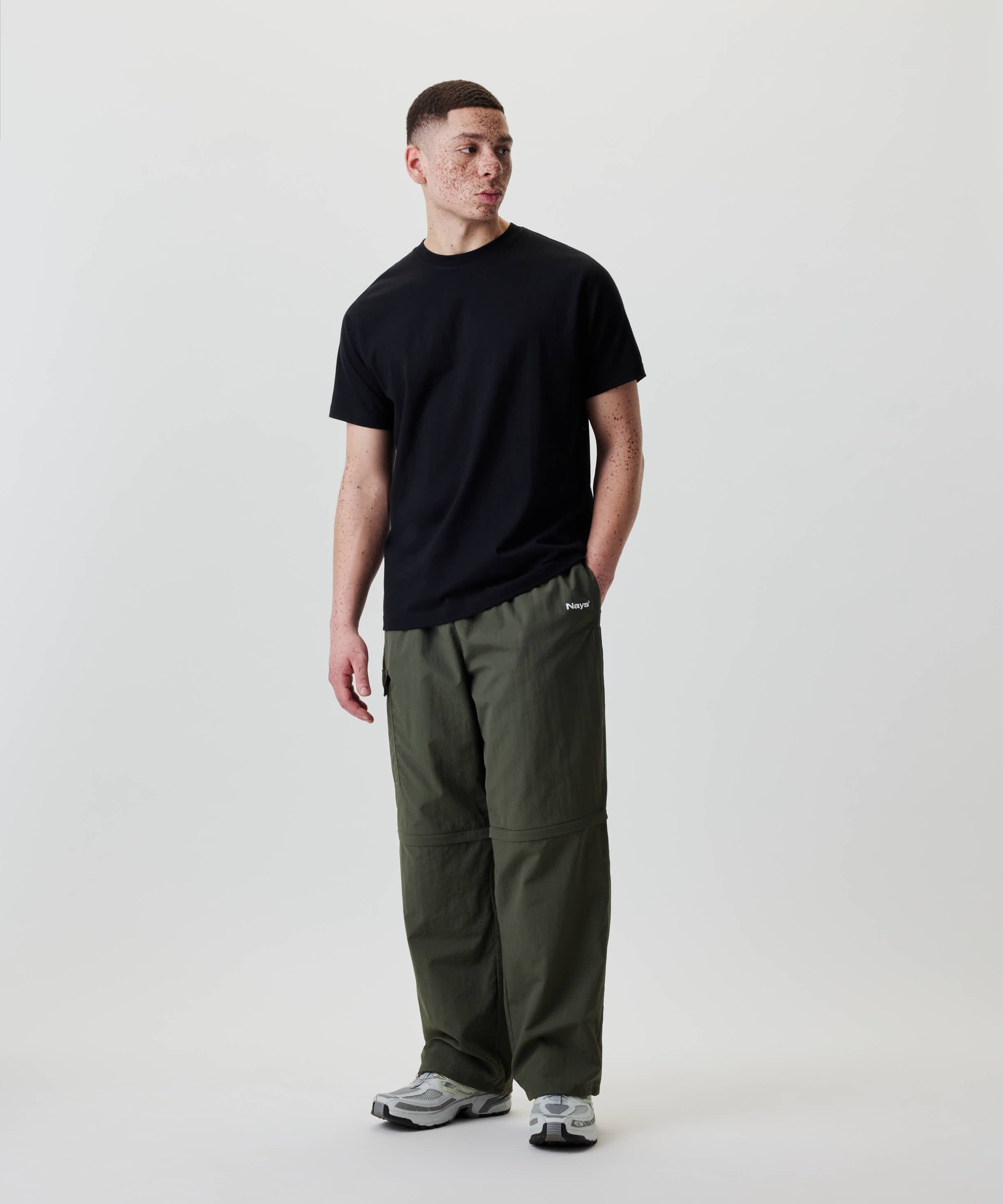 Junger Erwachsener trägt Nays Lightweight Zip Off Pants, wandelbar in Shorts, mit Logo am Oberschenkel und praktischen Reißverschlusstaschen, stehend gegen weißen Hintergrund.