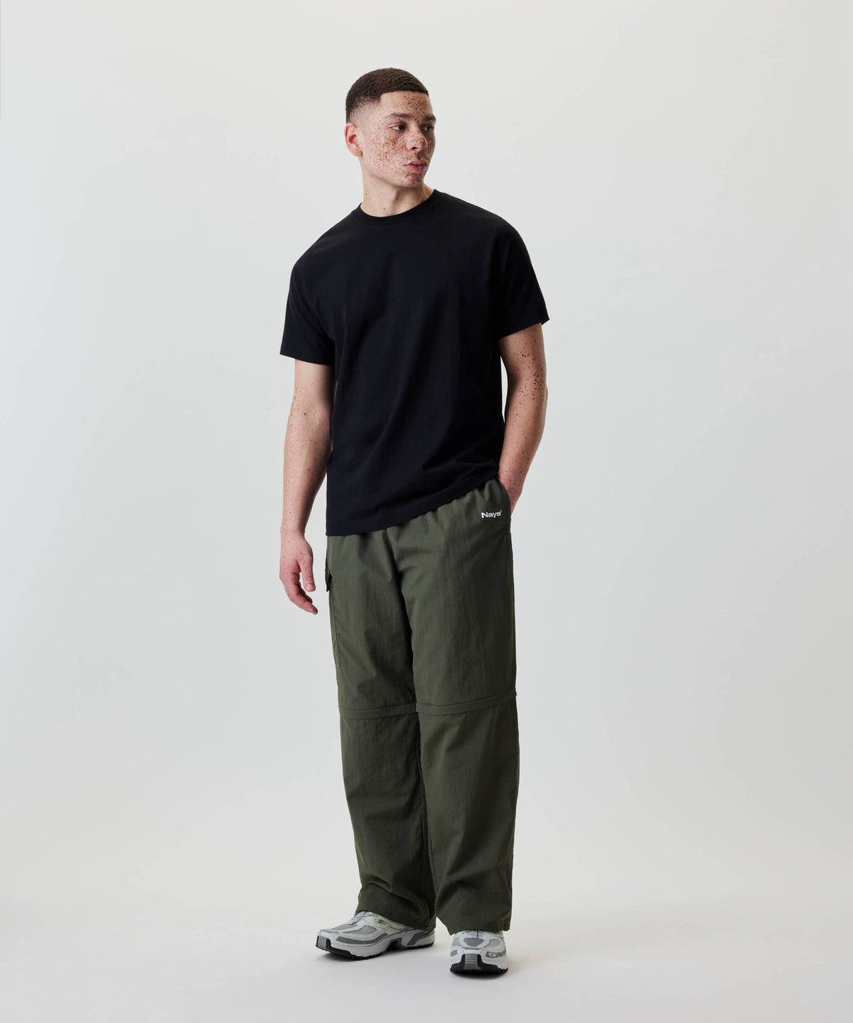 Junger Erwachsener trägt Nays Lightweight Zip Off Pants, wandelbar in Shorts, mit Logo am Oberschenkel und praktischen Reißverschlusstaschen, stehend gegen weißen Hintergrund.