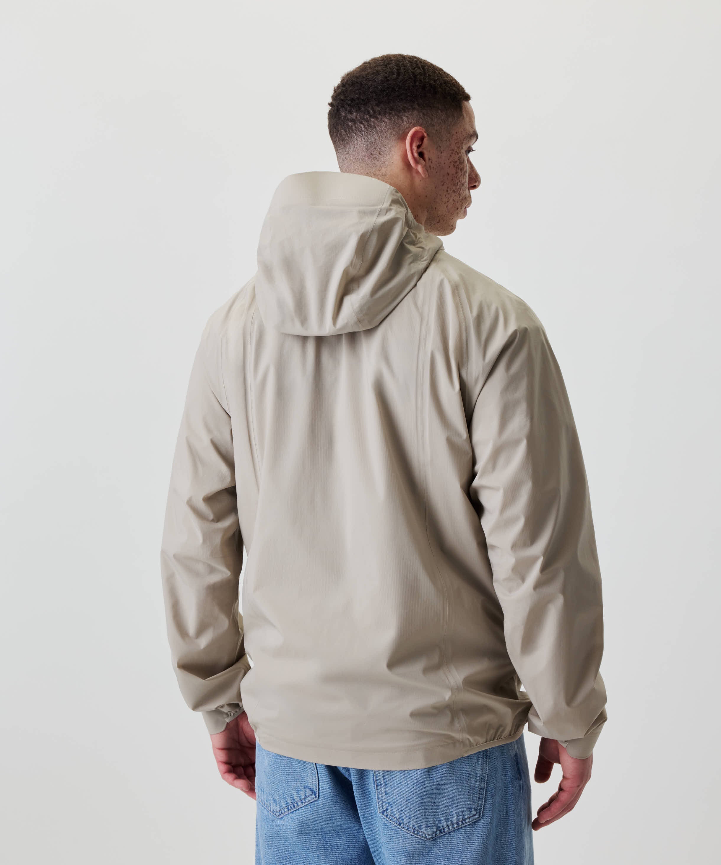 Rückansicht einer Person in Nays 2L Defend Mono Jacket, hellbeige, mit Kapuze und Raglanärmeln, kombiniert mit hellblauen Jeans, vor hellem Hintergrund.