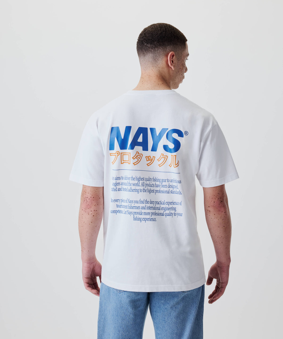 Eine Person trägt das Nays Pro Tackle International Tee, mit blauem NAYS-Schriftzug und orangefarbenen japanischen Zeichen auf dem Rücken, aus 100% Baumwolle.