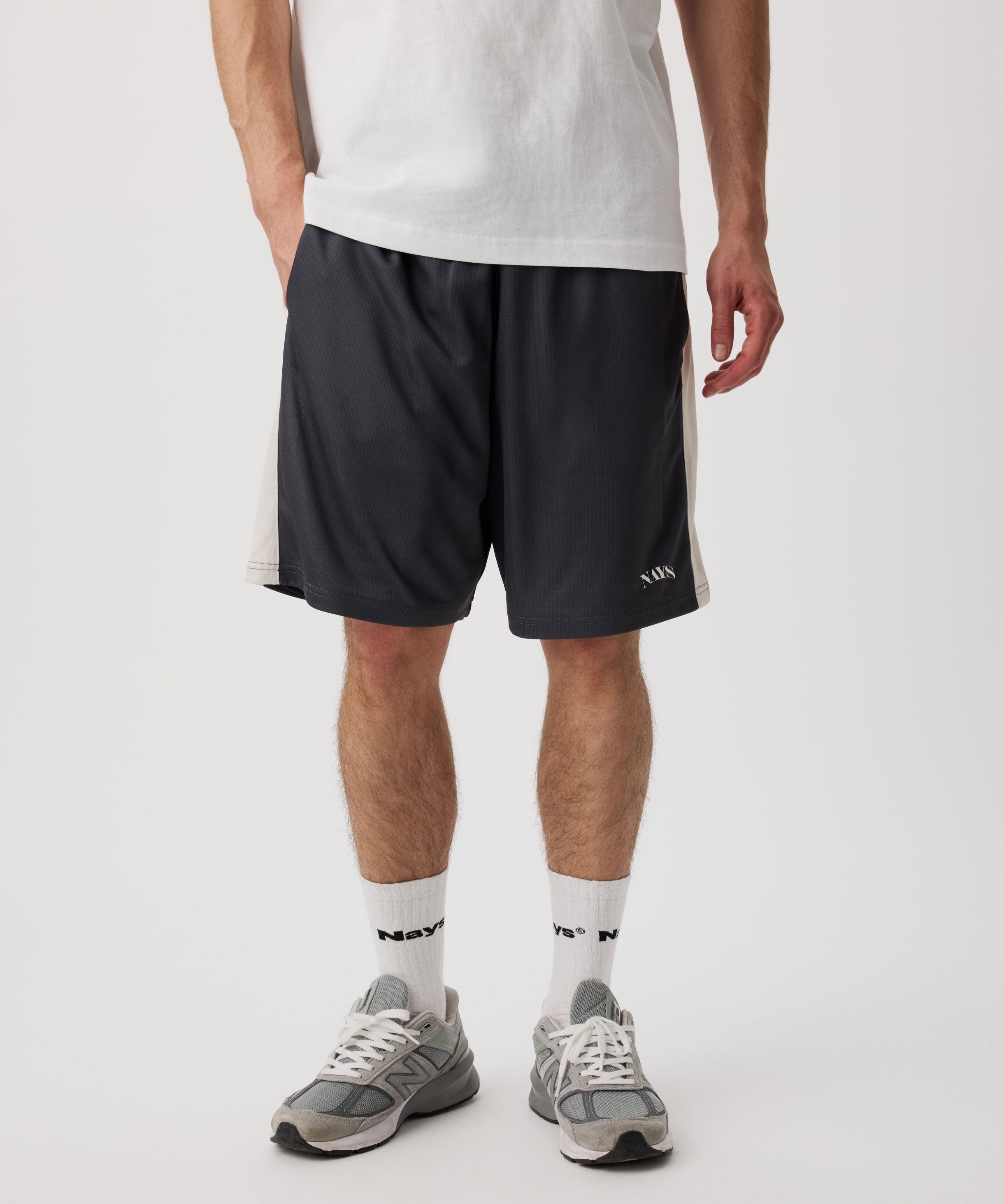 Das Model trägt eine blau-graue-kurze Hose mit weißen Applikationen an den Seiten. Dazu trägt er ein weißes T-Shirt, helle Socken und graue Turnschuhe.