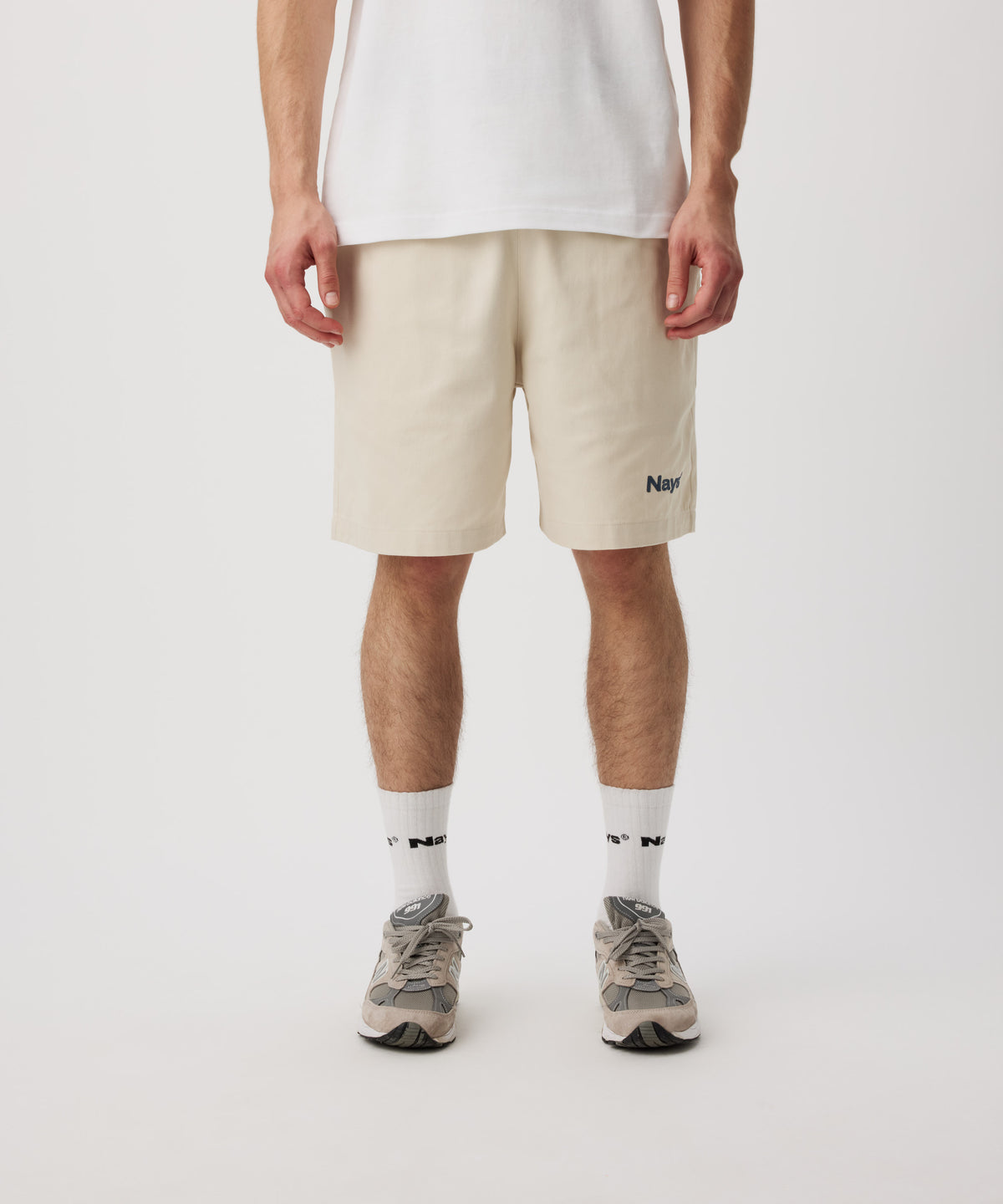 Ein Mann trägt ein weißes T-Shirt, eine beige Shorts mit einem kleinen, grünen Logo auf dem linken Bein. Dazu trägt er weiße Logo Socken und Sportschuhe.