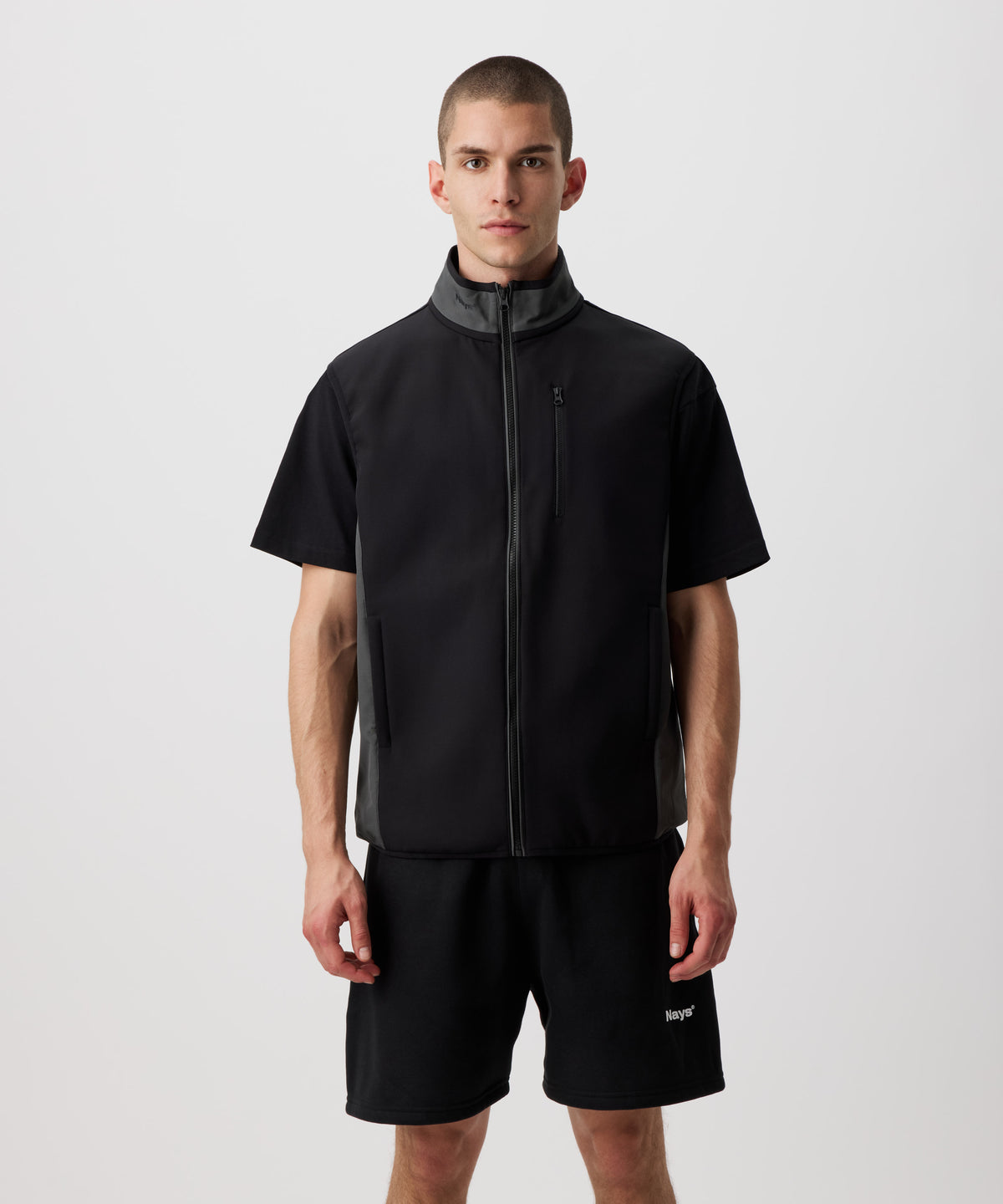Ein Mann trägt eine dünne, funktionelle Weste in schwarz. Der Reißverschluss, die Seiten und der Kragen sind farblich in grau abgesetzt. Dazu trägt er eine schwarze Shorts.
