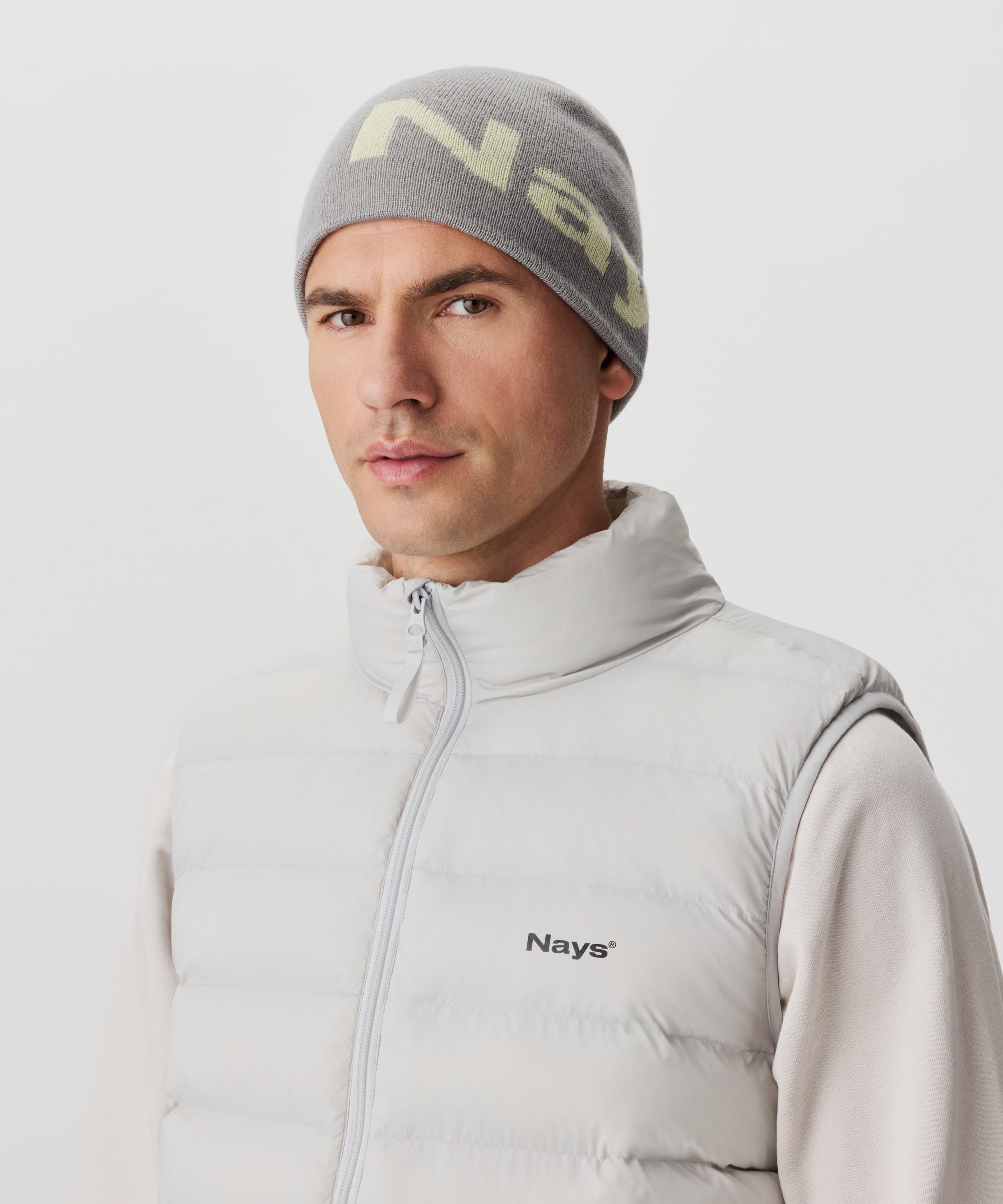Frontansicht Mann mit grauer Strickmütze und farblich abgesetztem Nays-Logo. Er trägt einen beige-farbigen Pullover und eine hell-graue Weste mit schwarzem Logo auf der linken Brust.