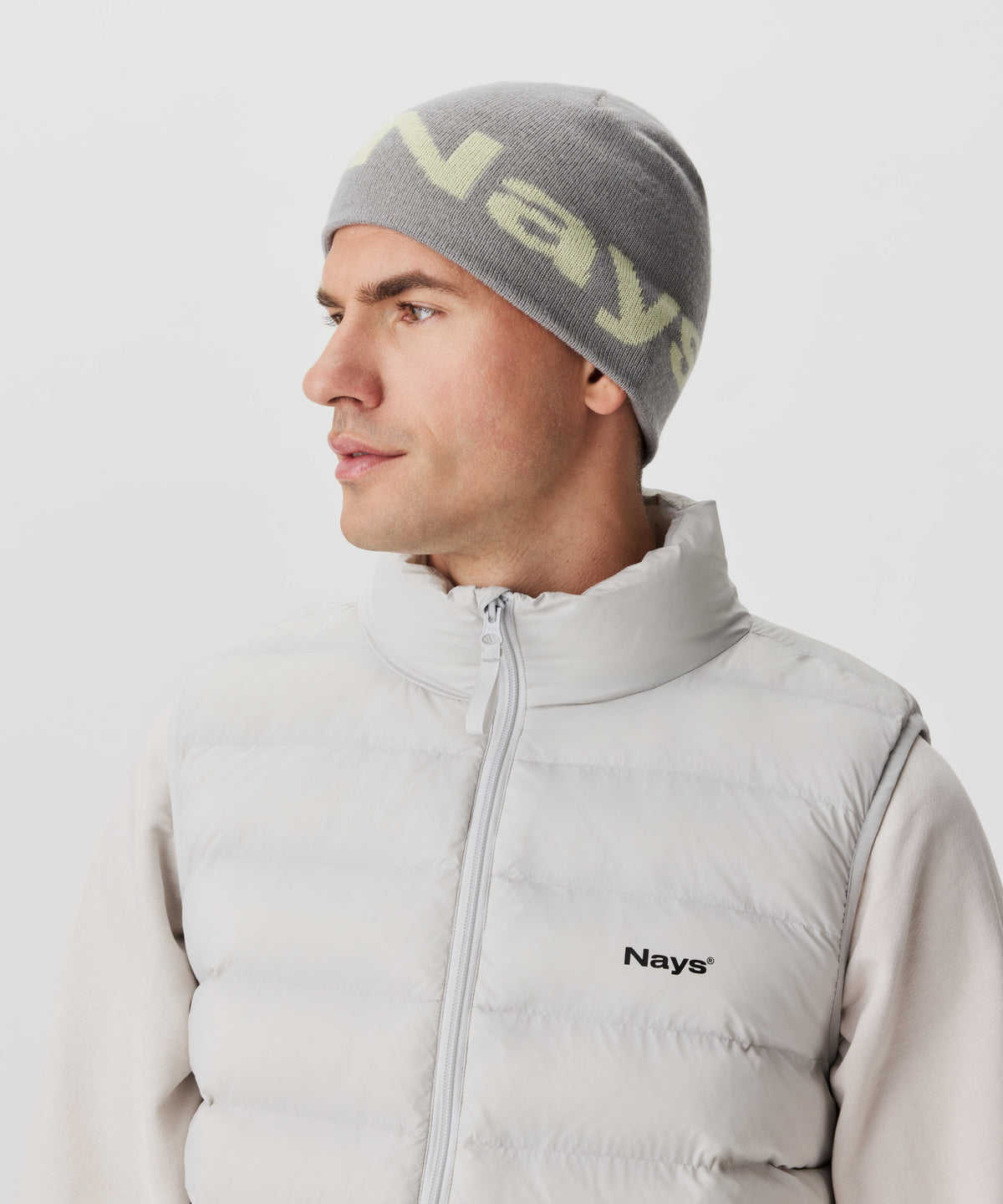 Ansicht im Halbseitenprofil Mann mit grauer Strickmütze und farblich abgesetztem Nays-Logo. Er trägt einen beige-farbigen Pullover und eine hell-graue Weste mit schwarzem Logo auf der linken Brust.