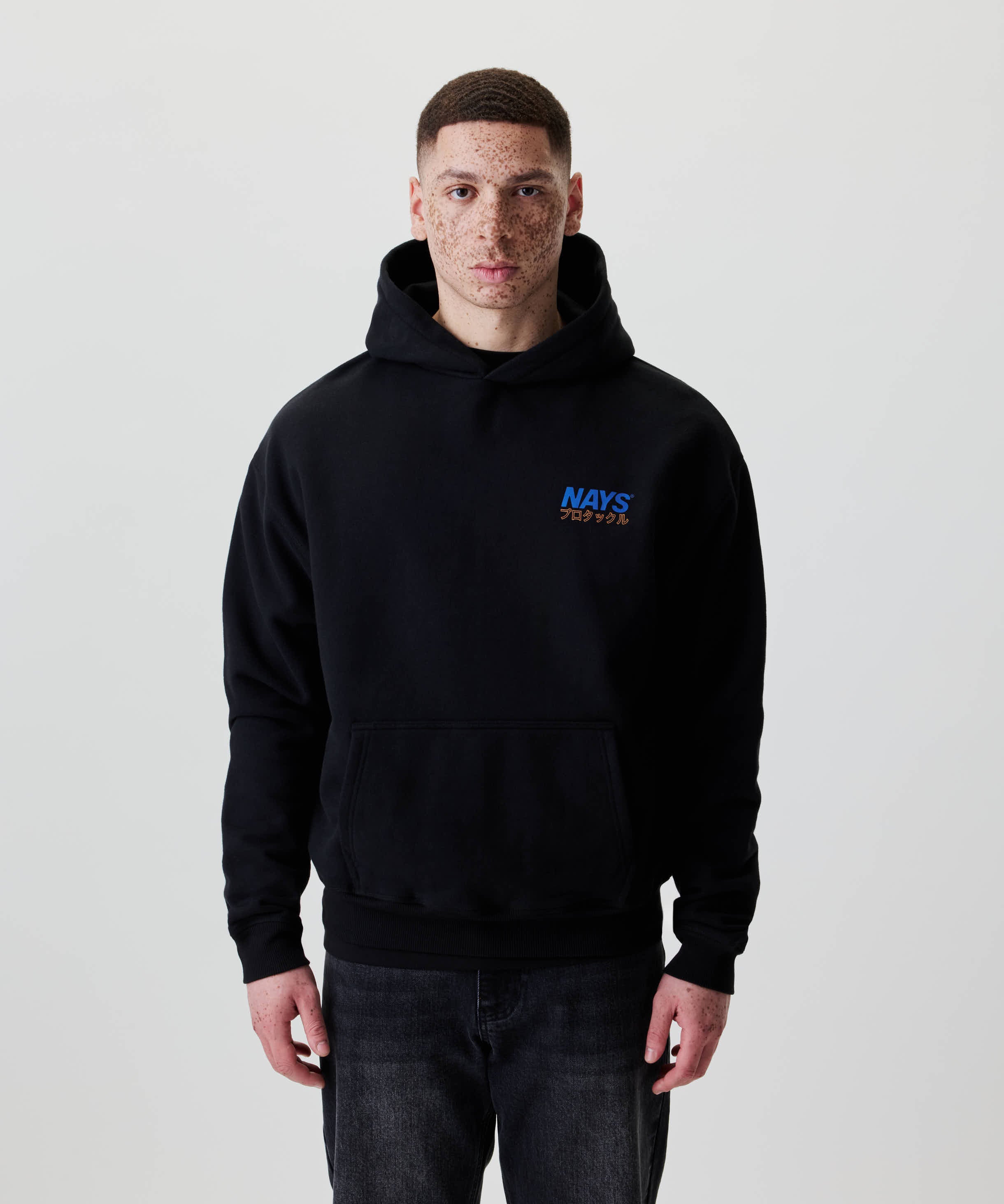 Junger Mann trägt Nays Pro Tackle International Hoodie mit kleinem Logo und Kängurutasche, kombiniert mit dunklen Jeans. Ideal für Alltag und Freizeit.