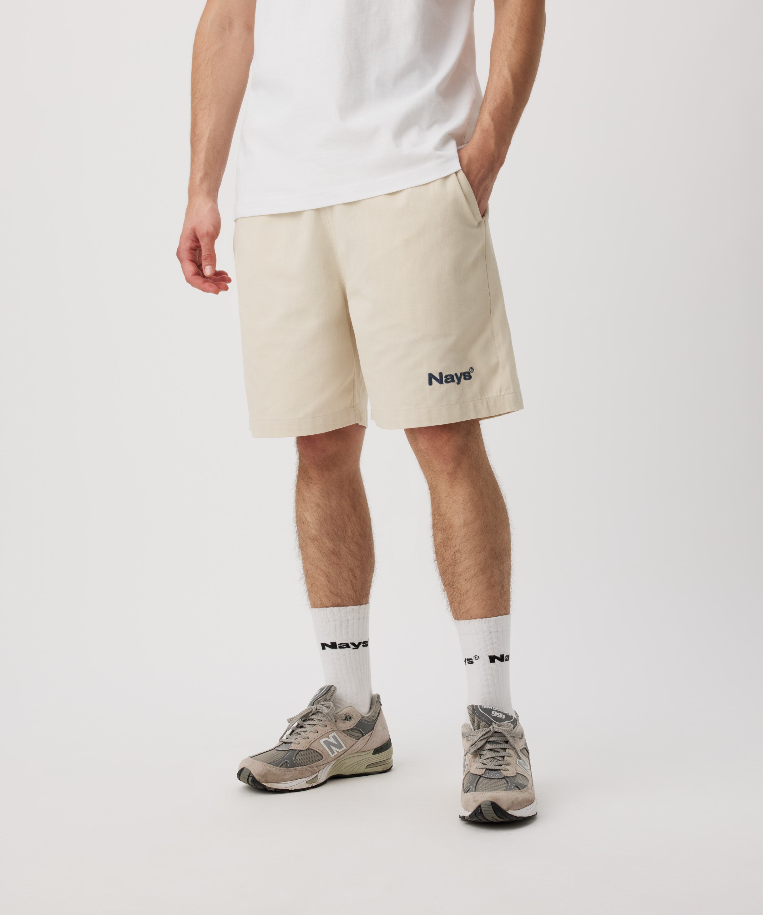 Ein Mann steht schräg zur Kamera. Er trägt ein weißes T-Shirt, eine beige Shorts mit einem kleinen, grünen Logo auf dem linken Bein. Dazu trägt er weiße Logo Socken und Sportschuhe.