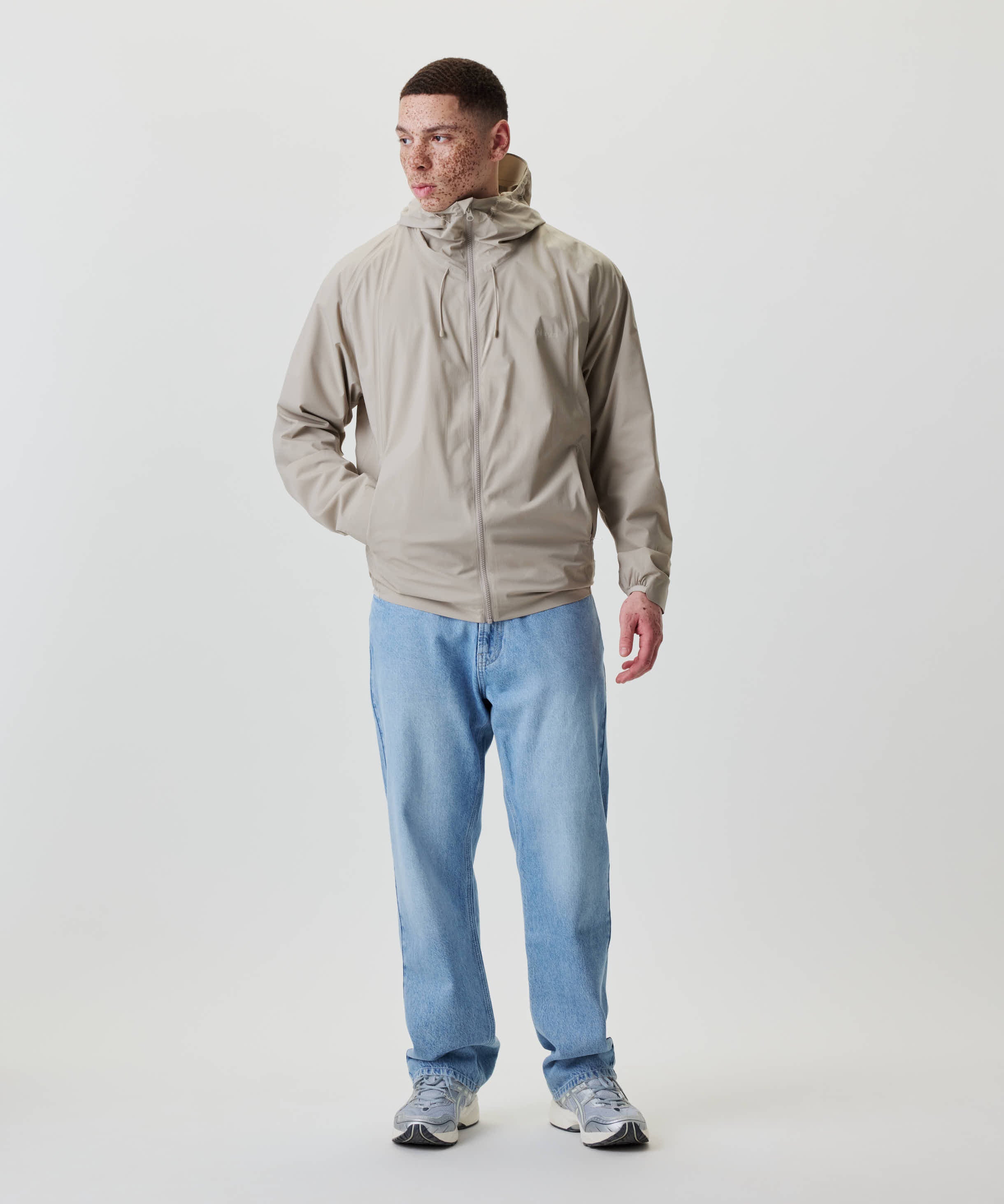 Ein junger Mann trägt die Nays 2L Defend Mono Jacket mit Kapuze, sportlichen Raglanärmeln und elastischen Bündchen, kombiniert mit Jeans und Sneakers.