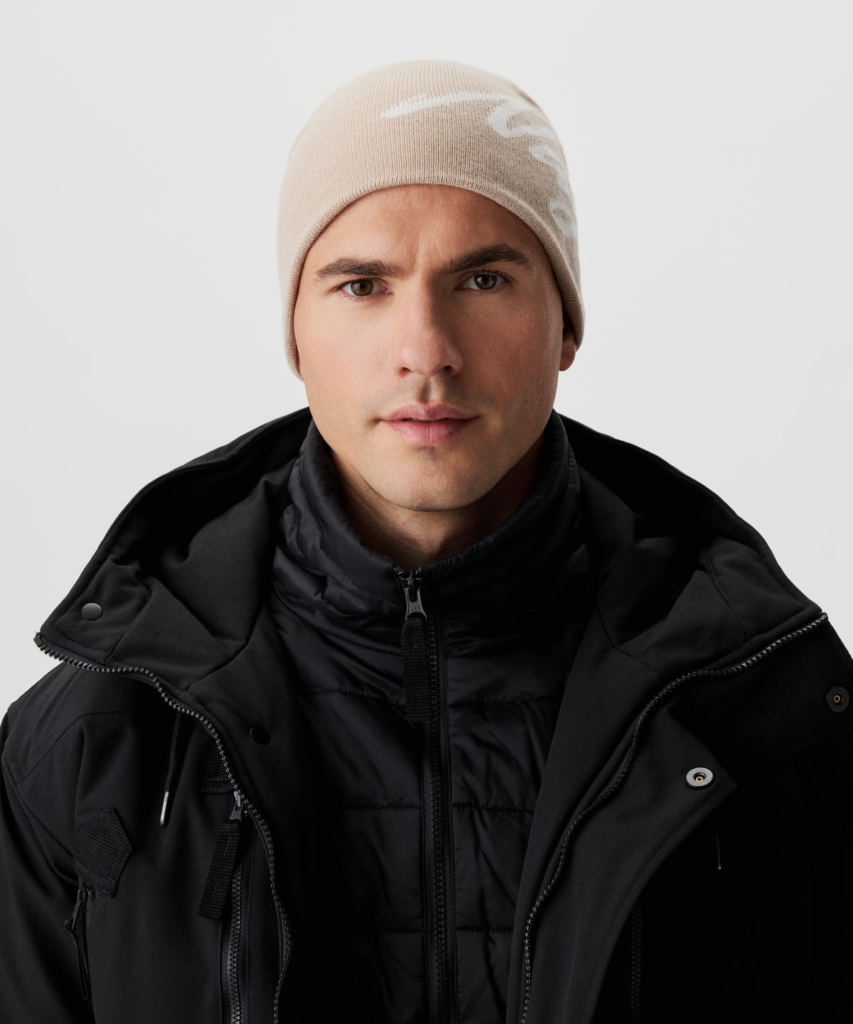 Frontale Ansicht Mann  in schwarzer Jacke und beige-farbiger Strickmütze mit weißem Nays-Logo-Schriftzug.