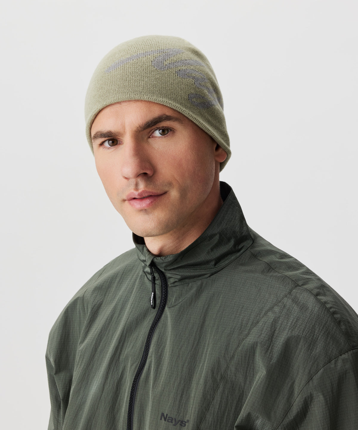 Frontale ansicht Mann in grüner Jacke und grüner Strickmütze mit grauen Nays-Logo-Schriftzug.
