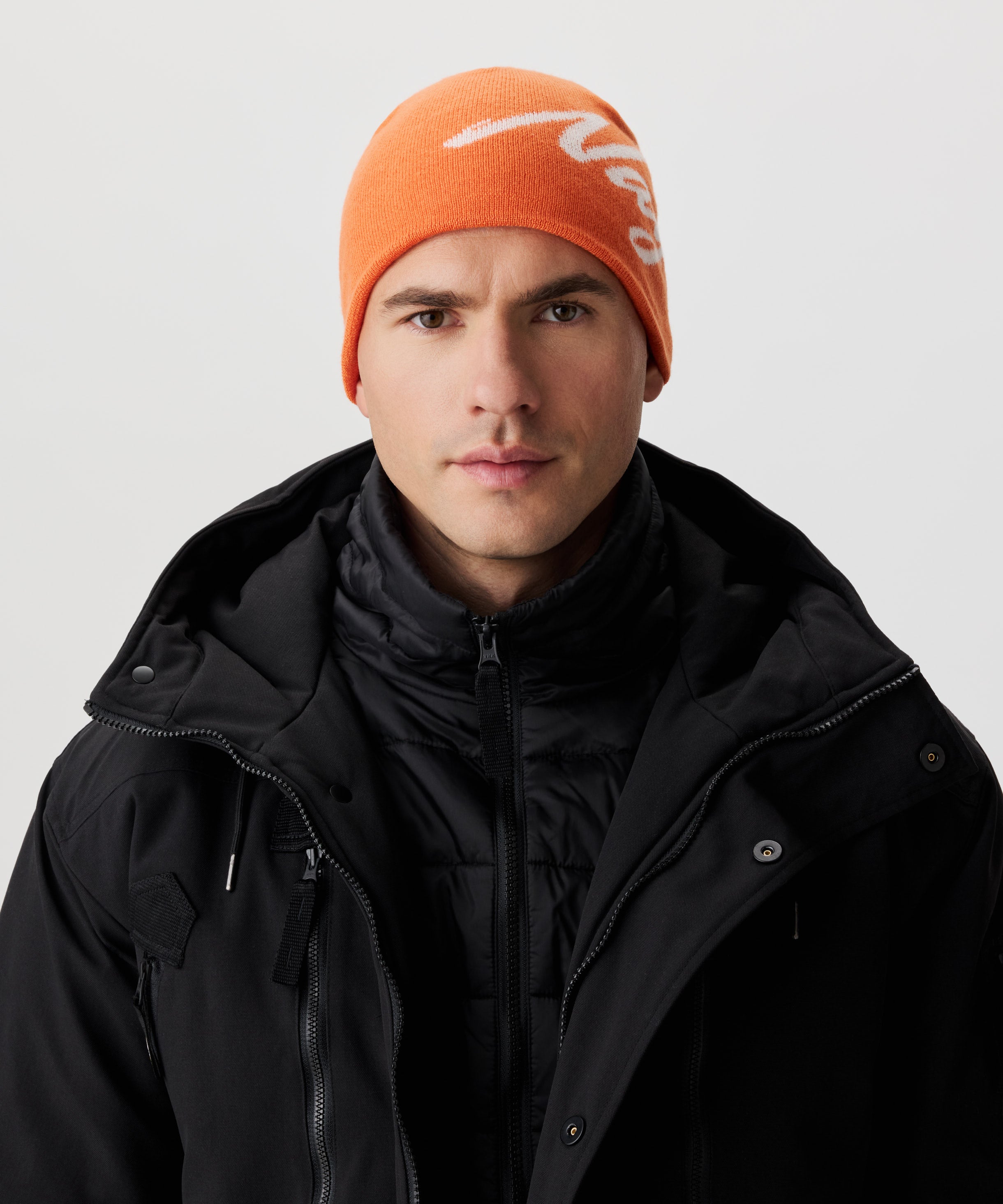 Frontale Ansicht Mann  in schwarzer Jacke und orange-farbiger Strickmütze mit weißem Nays-Logo-Schriftzug.