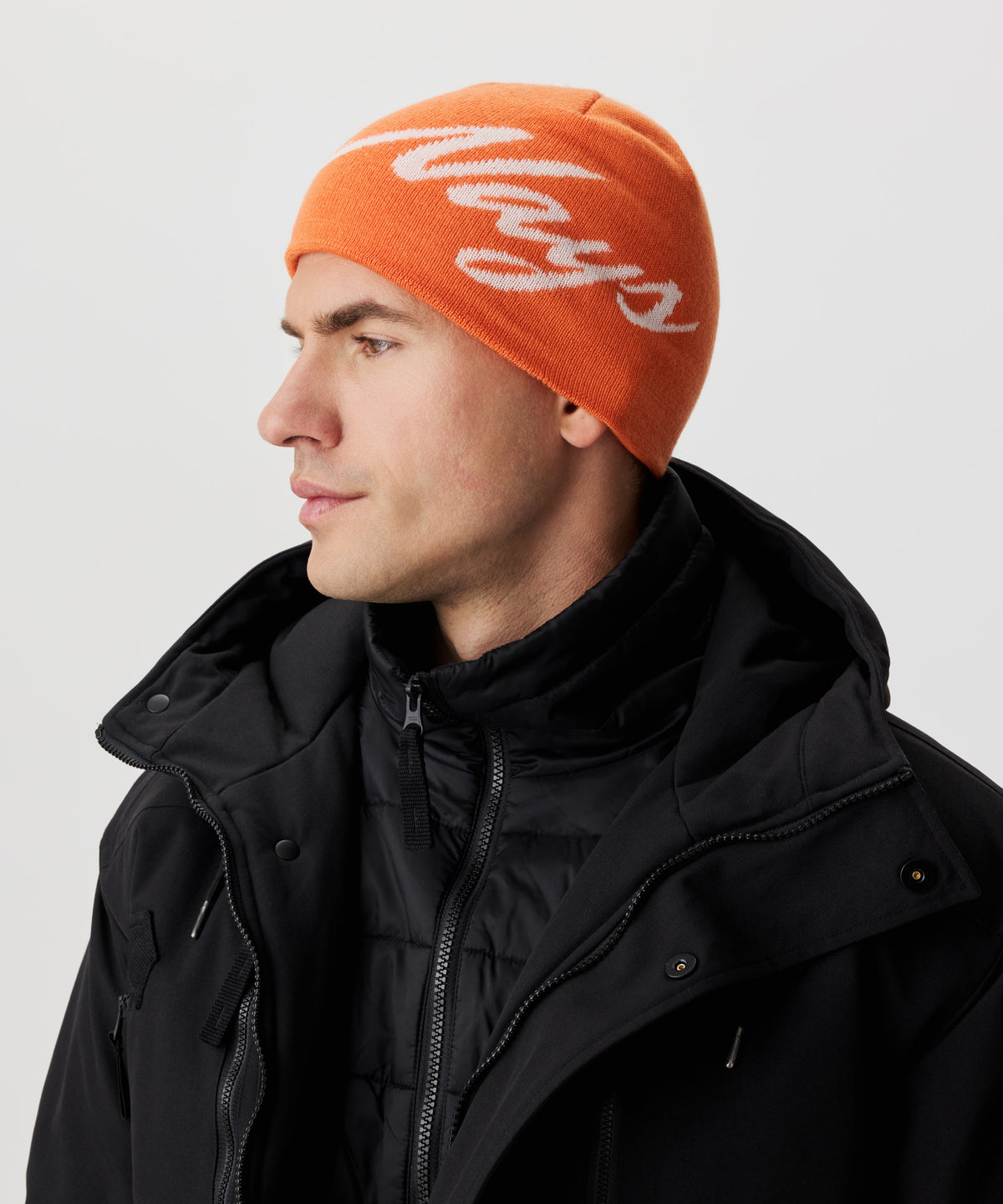 Seitliche Ansicht Mann  in schwarzer Jacke und orange-farbiger Strickmütze mit weißem Nays-Logo-Schriftzug.