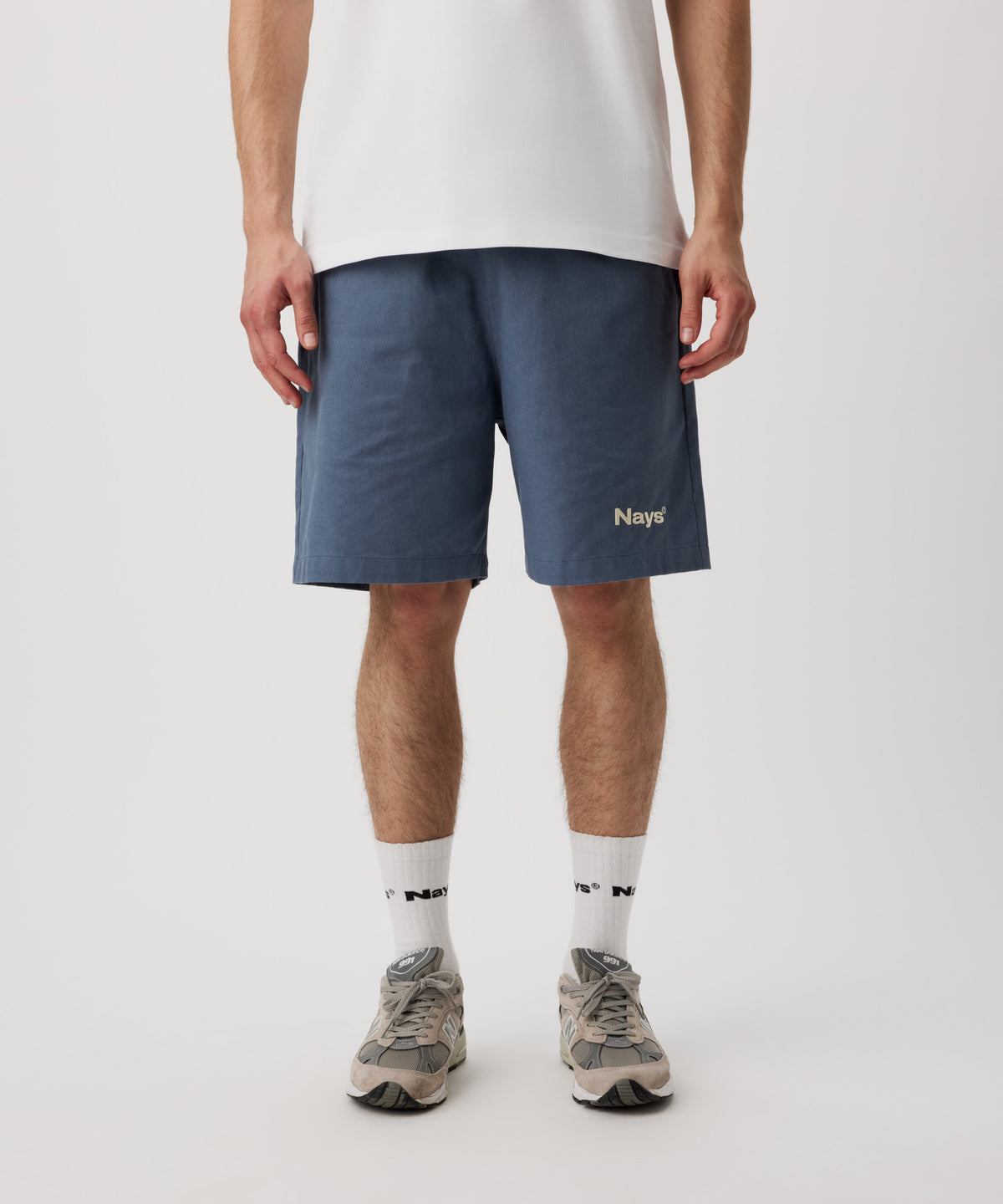 Ein Mann trägt ein weißes T-Shirt, eine blaue Shorts mit einem kleinen, weißen Logo auf dem linken Bein. Dazu trägt er weiße Logo Socken und graue Sportschuhe.