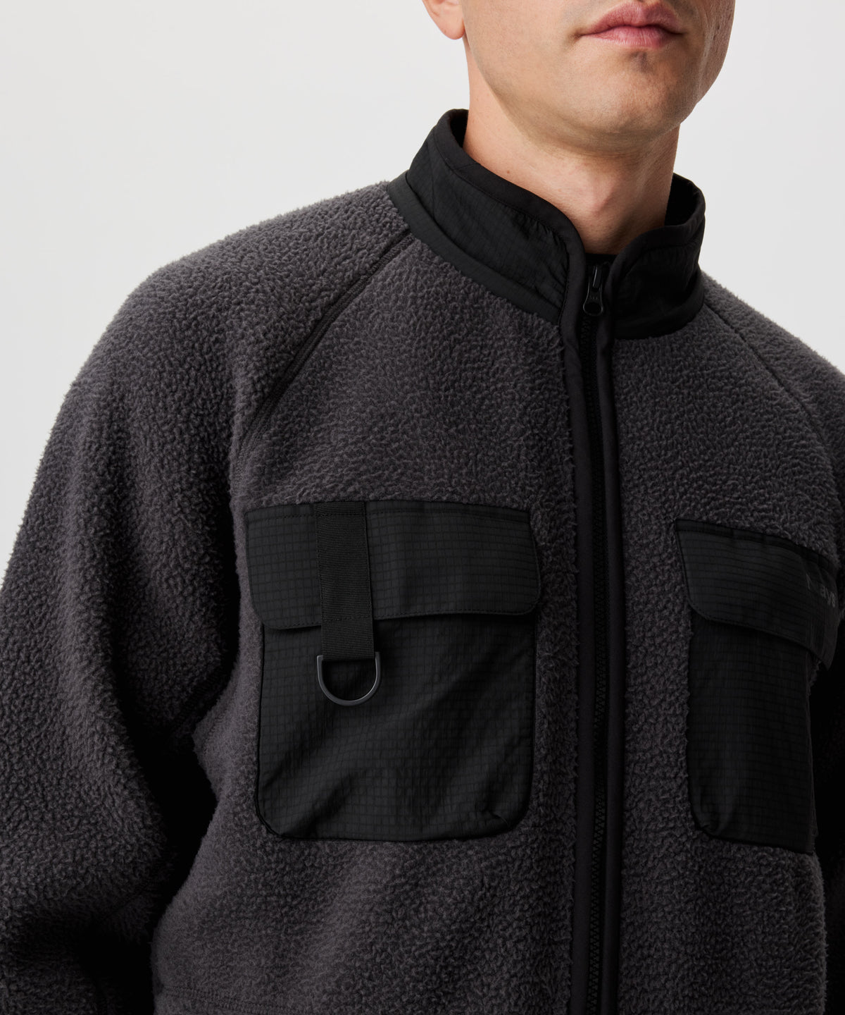Ansicht von einem Mann in dunkel-grauer Fleece-Jacke mit abgesetzten Kragen und Brusttaschen im schwarzen Ripstop. Die rechte Brusttasche verfügt über einen scwharzen D-Ring.