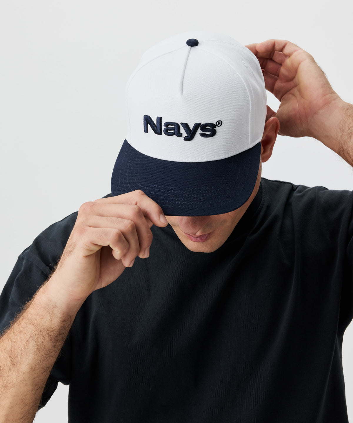 Ein Mann setzt eine Cap auf. Die Cap ist weiß mit einem blauen Schirm und blauem Nays-Logo Strick.