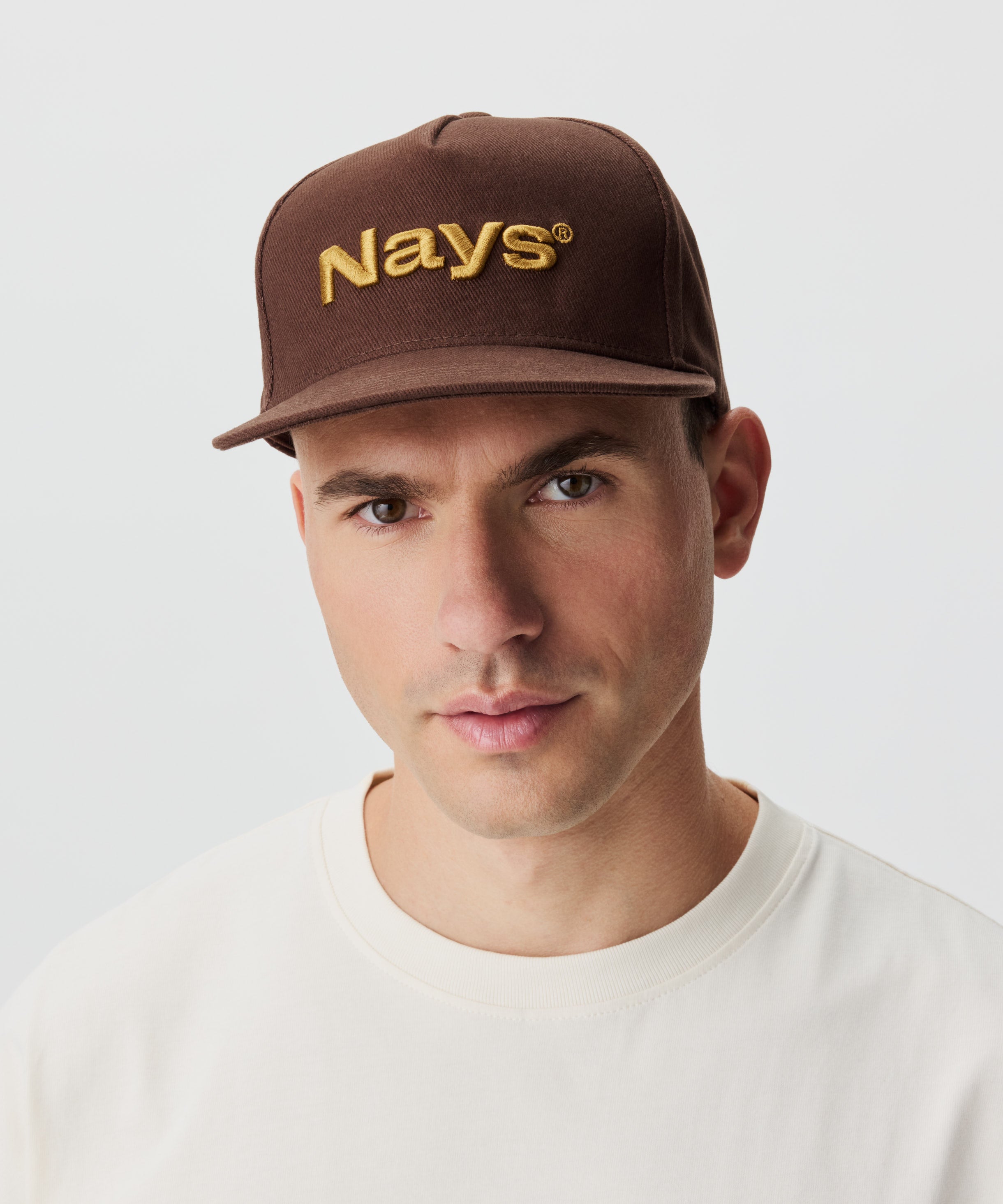 Frontansicht Mann mit Cap. Die Cap ist braun mit einem gold-gelbem Nays-Logo Strick.