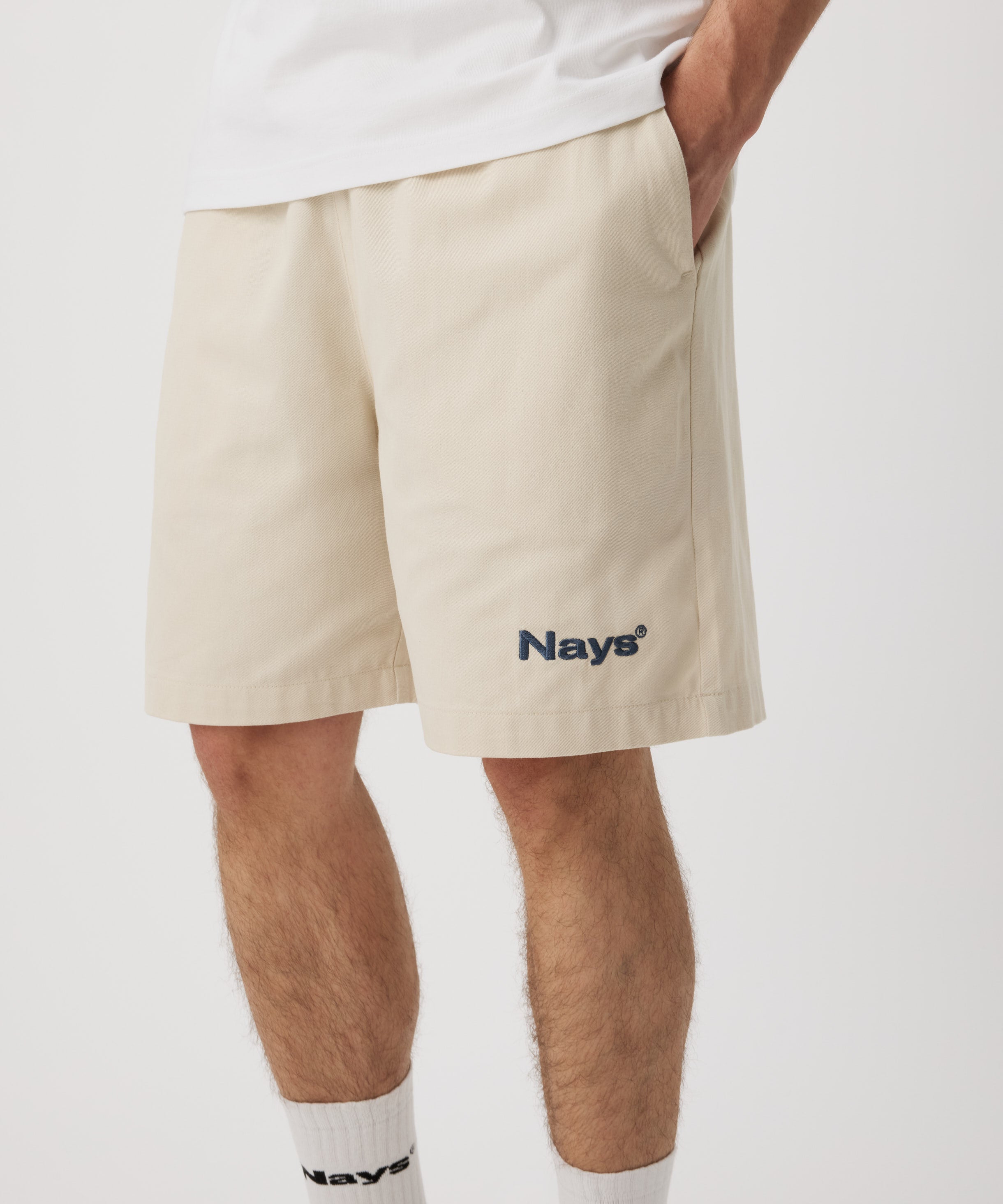 Ein Mann trägt eine beige Shorts mit einem dunkel-grünen "Nays"-Logo auf dem linken Bein und hat seine linke Hand in der Tasche. Dazu trägt er ein weißes T-Shirt und eine weiße Socken.