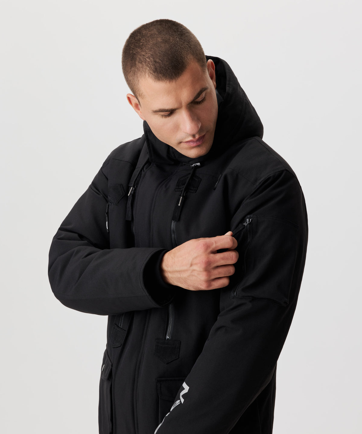 Man greift in eine Tasche auf dem linken Oberarm einer schwarzen Winterjacke mit Kapuze.