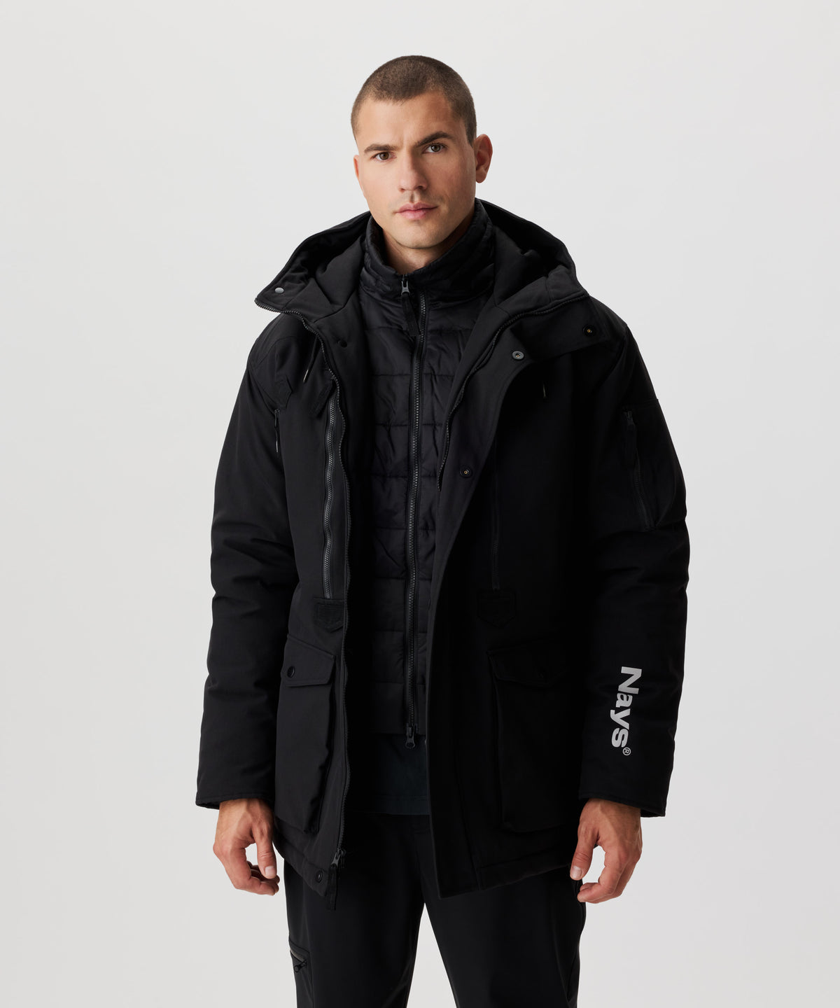 Frontansicht Mann im schwarzen Winter-Parka. Die äußere Jacke ist geöffnet und man sieht die darunter liegende, abgesteppte Daunenjacke.