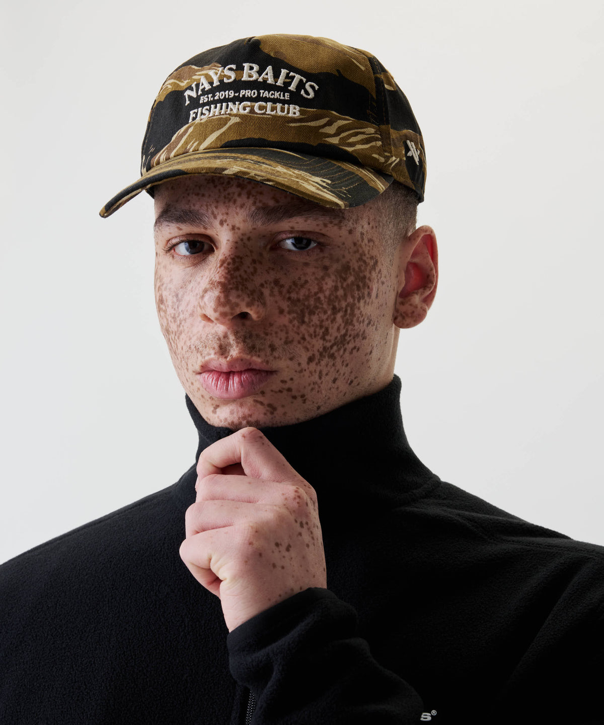 Nahaufnahme einer Person mit Sommersprossen, die die Nays Fishing Club Cap mit Camo-Print und off-white Stickerei trägt, hält den Kragen ihres schwarzen Oberteils.
