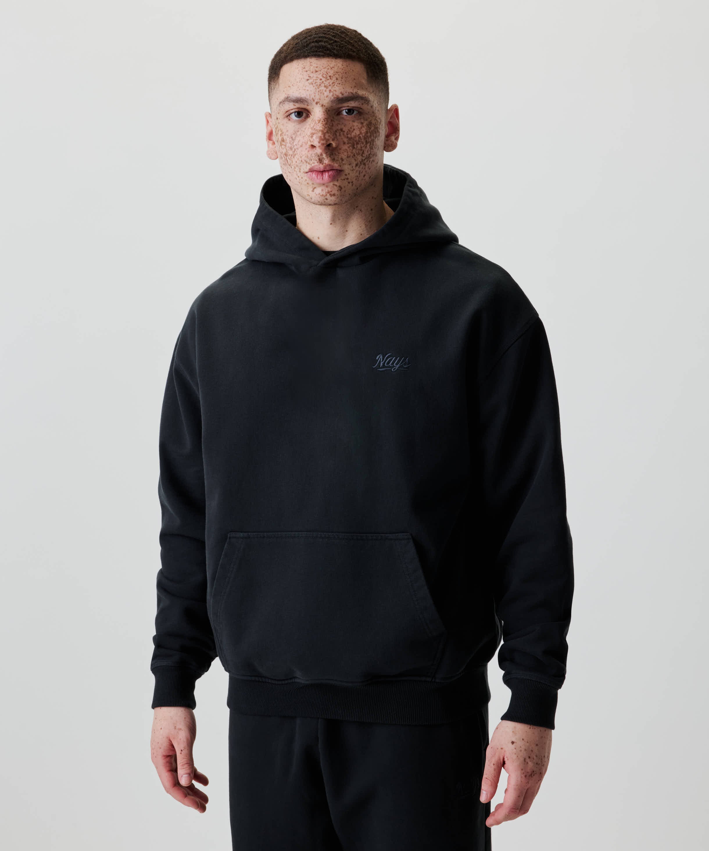 Junger Mann trägt das Nays New Script Hoodie in Schwarz mit dezentem Logo auf der Brust. Der Hoodie bietet einen lässigen und zeitlosen Look.